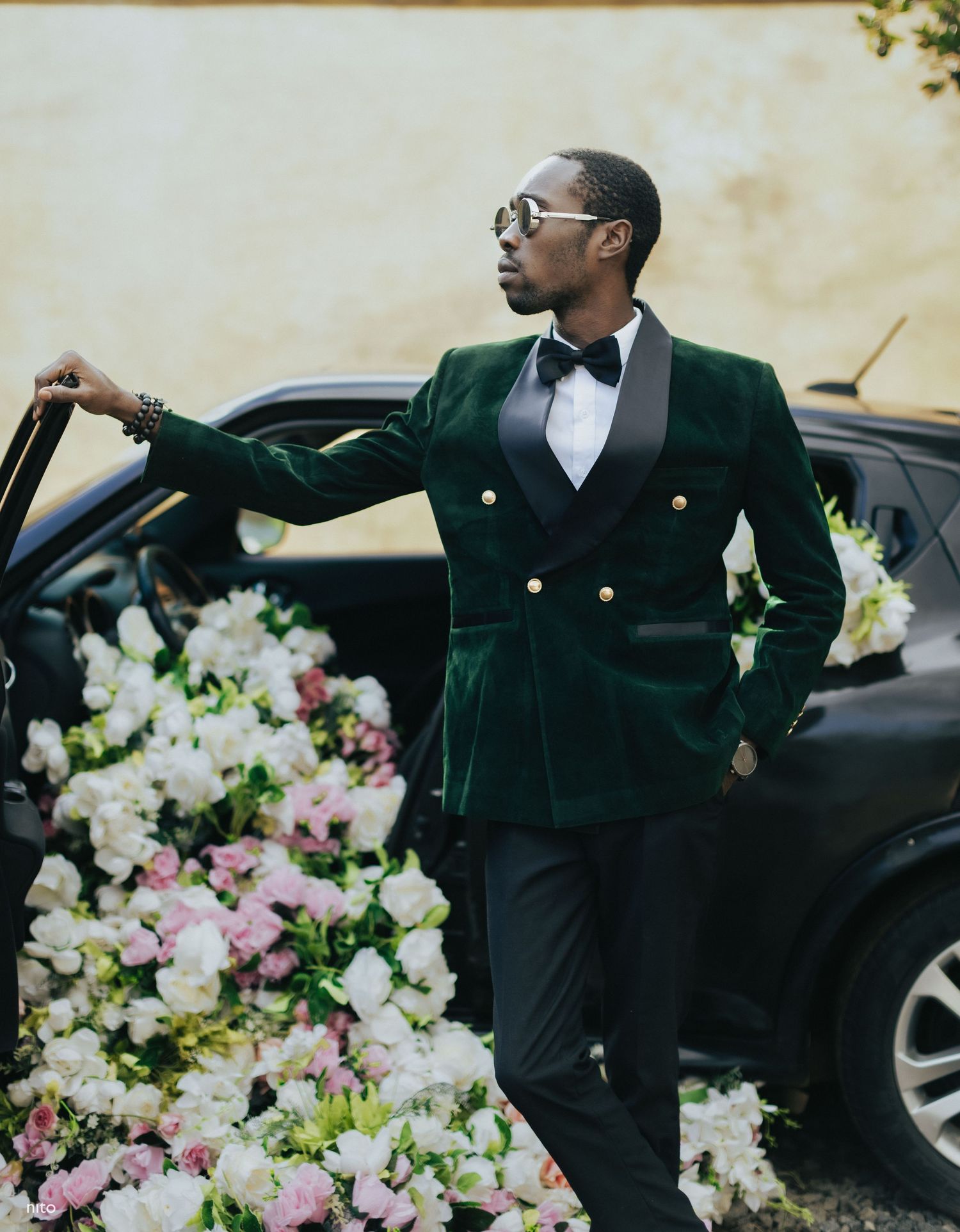 Person in green velvet suit jacket posing confidently next to car with floral arrangements.