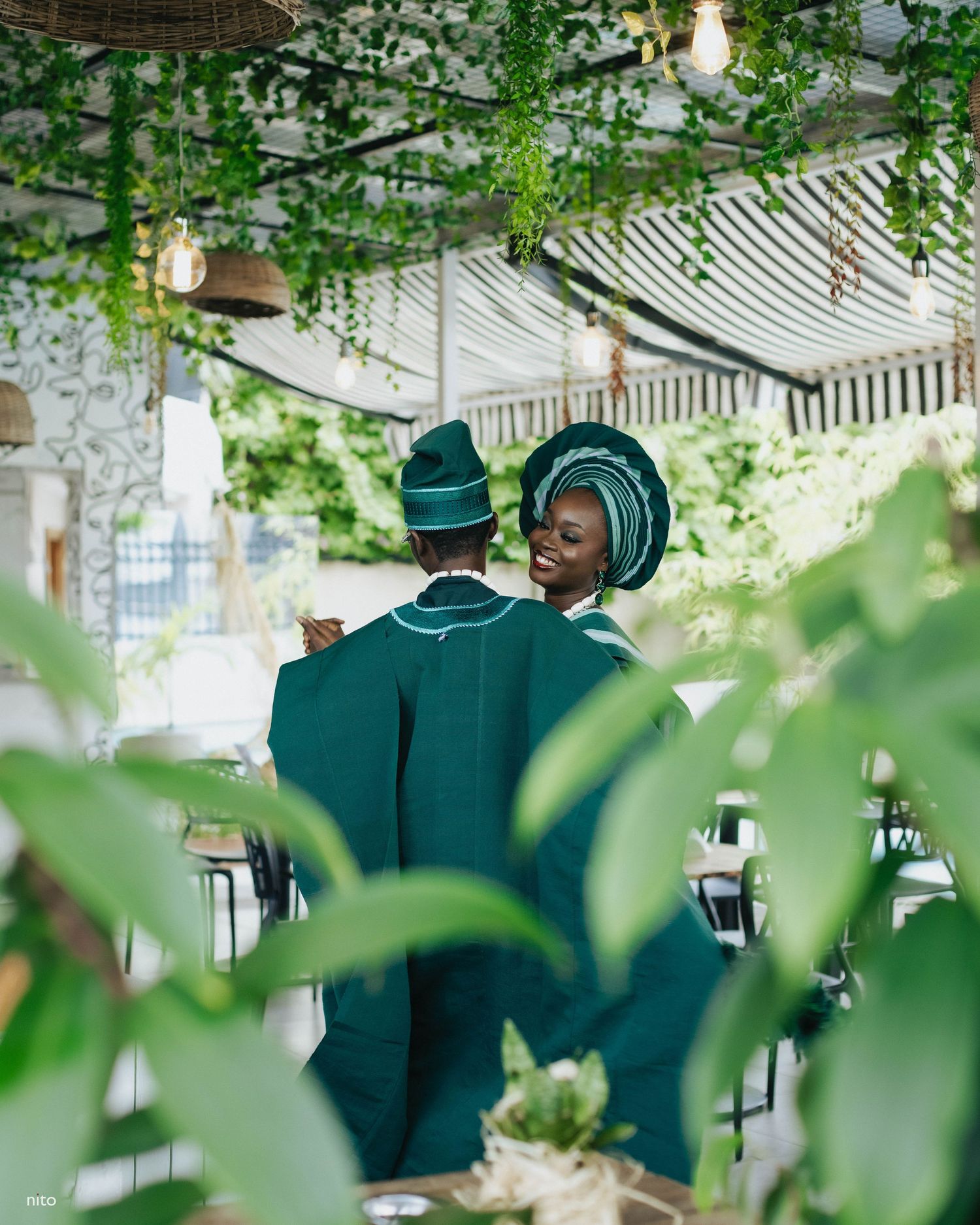 Traditional wedding couple in emerald green attire photographed through leafy decor.