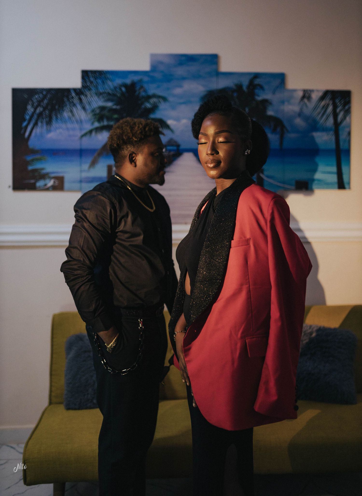 Two people posing indoors with a tropical beach backdrop painting behind them.