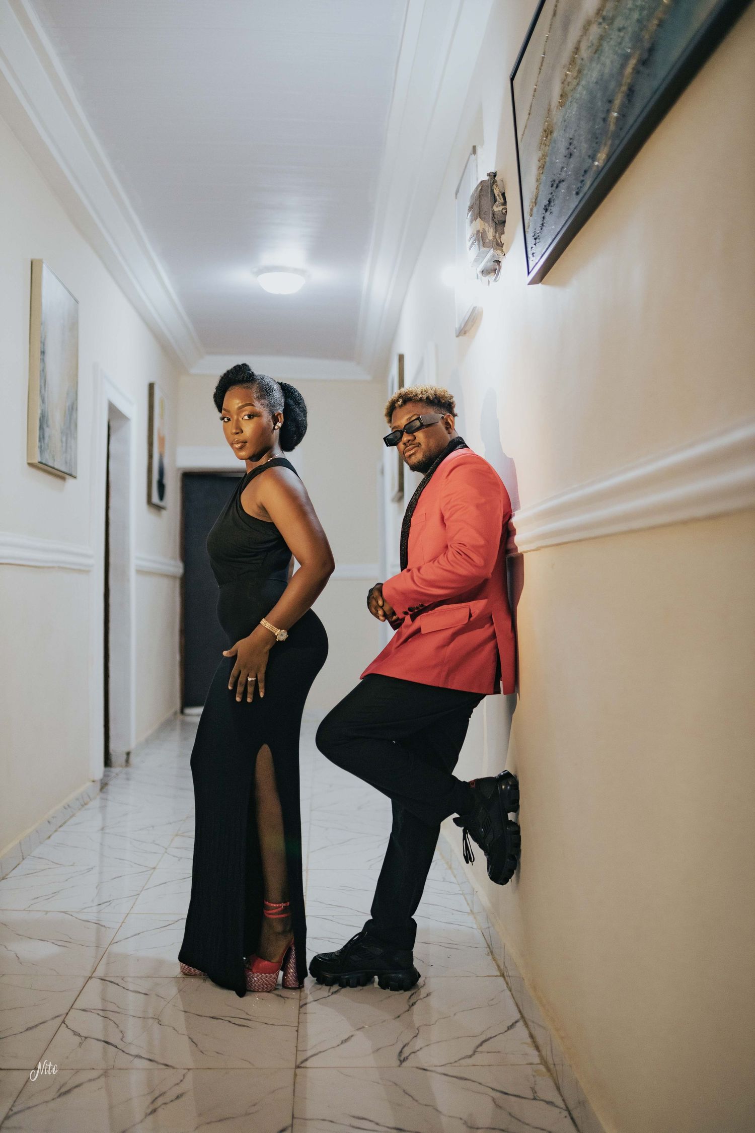 A stylish couple poses in a hallway, one in black dress and one in coral blazer.