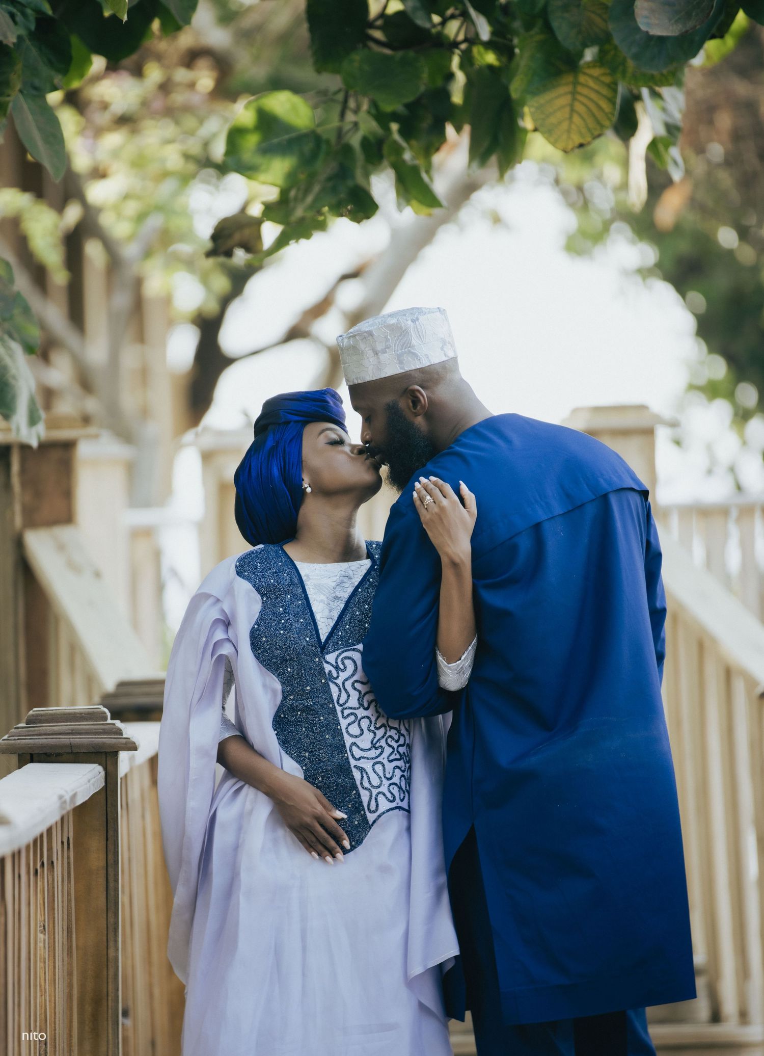 A couple in blue and white traditional dress sharing a tender moment by a wooden fence.