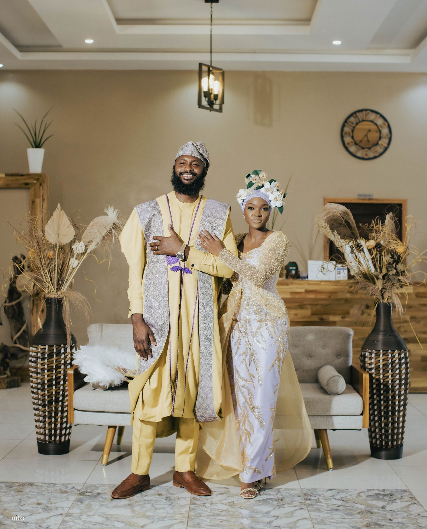 A couple wearing coordinated cream and white traditional outfits pose in their modern living space.