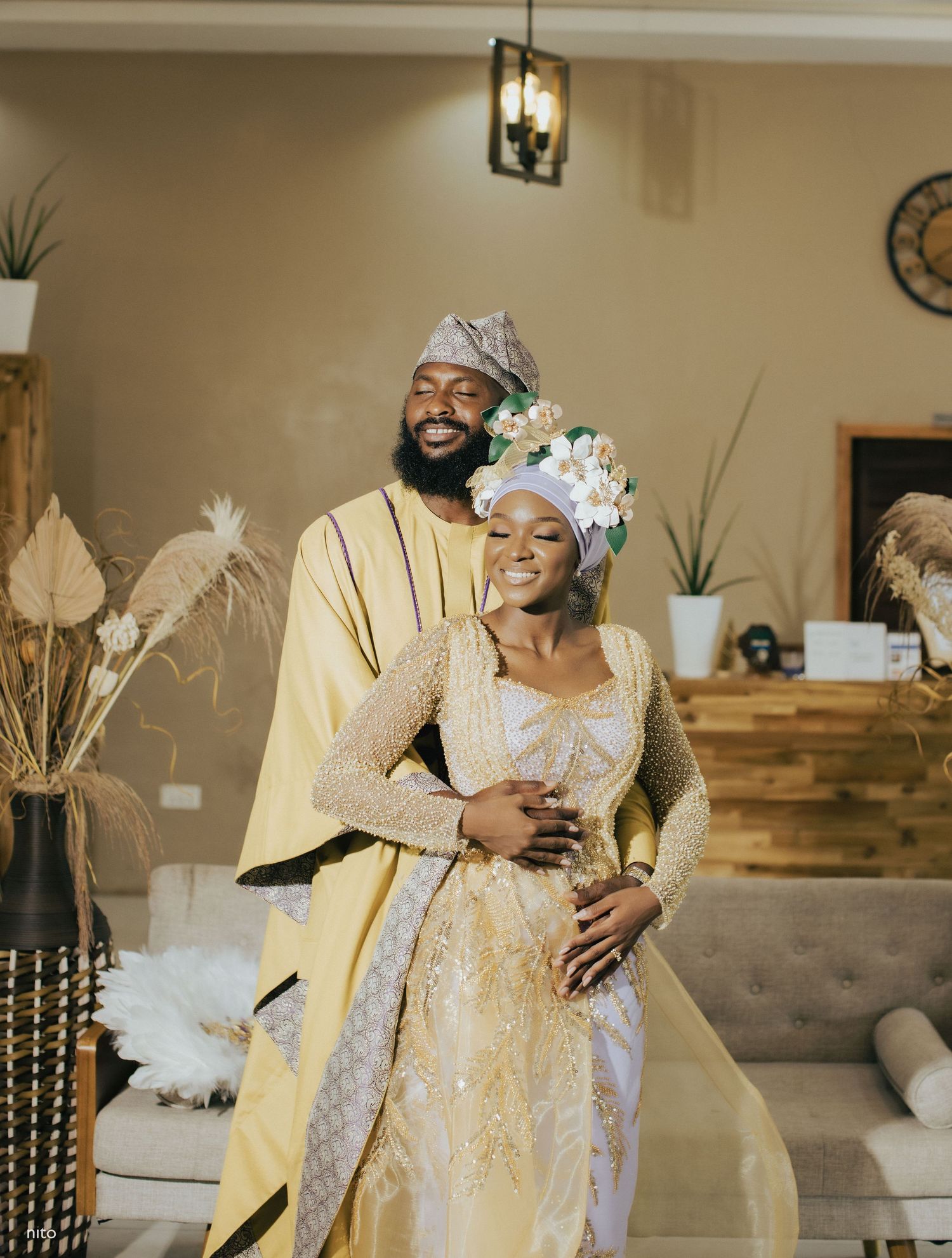 A couple in cream and white traditional attire embracing in their decorated living room.