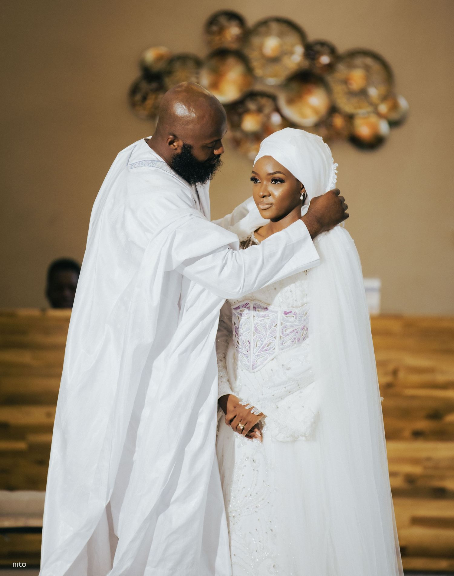 A couple in white traditional wedding attire sharing an intimate moment indoors.