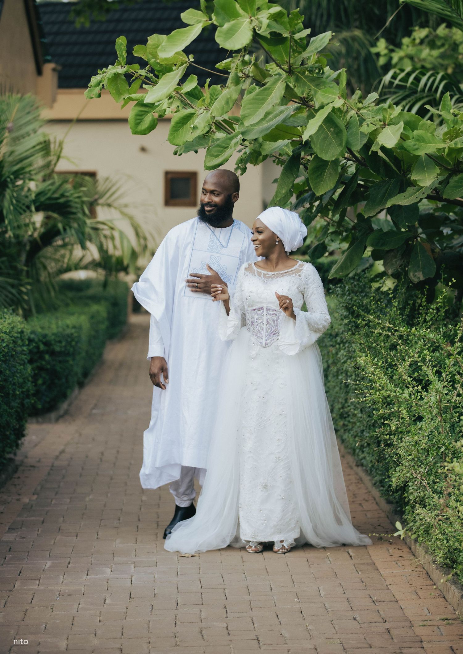 A couple in elegant white traditional attire walking along a garden pathway.