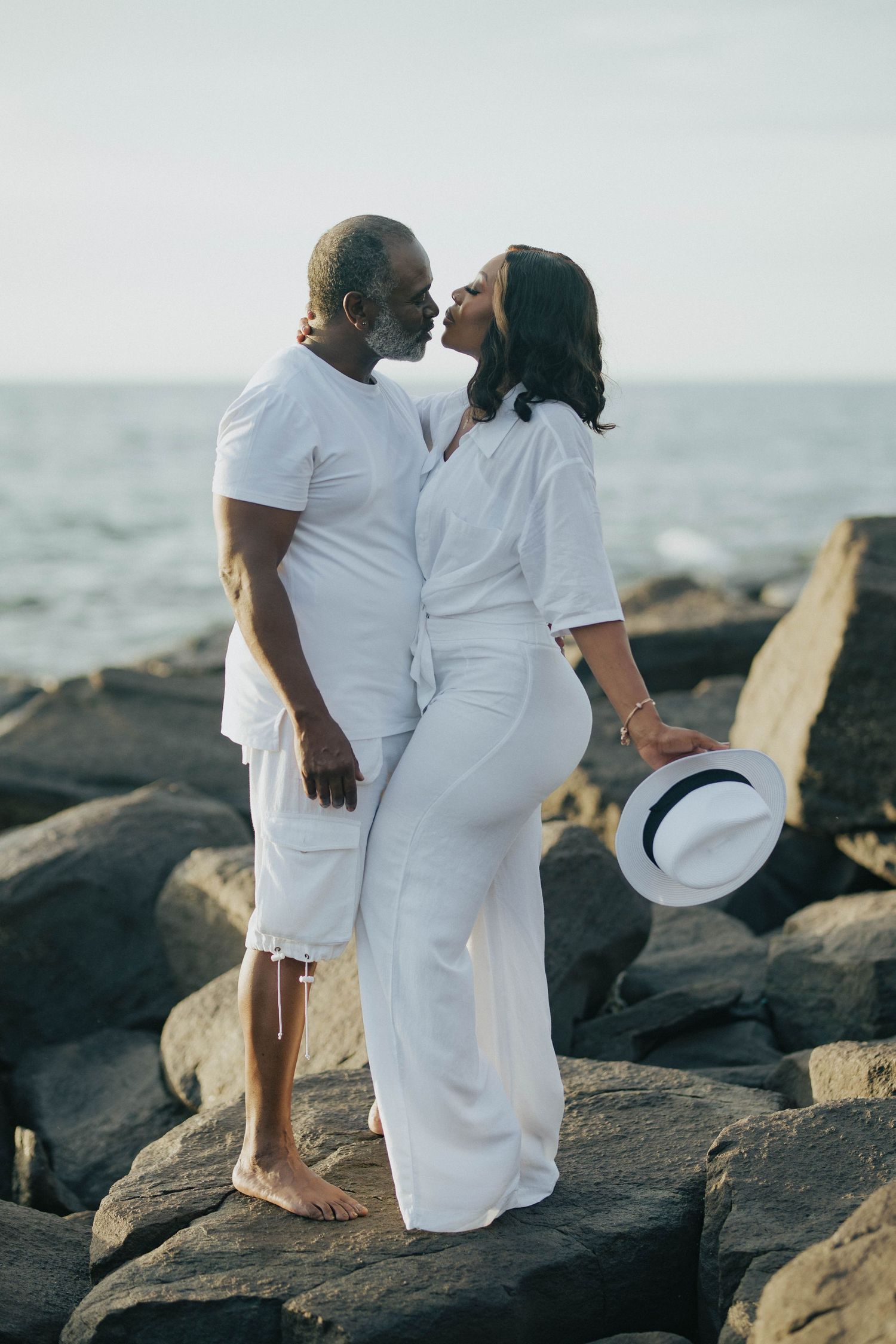 Romantic couple in white outfits sharing an intimate moment on rocky beach at sunset.