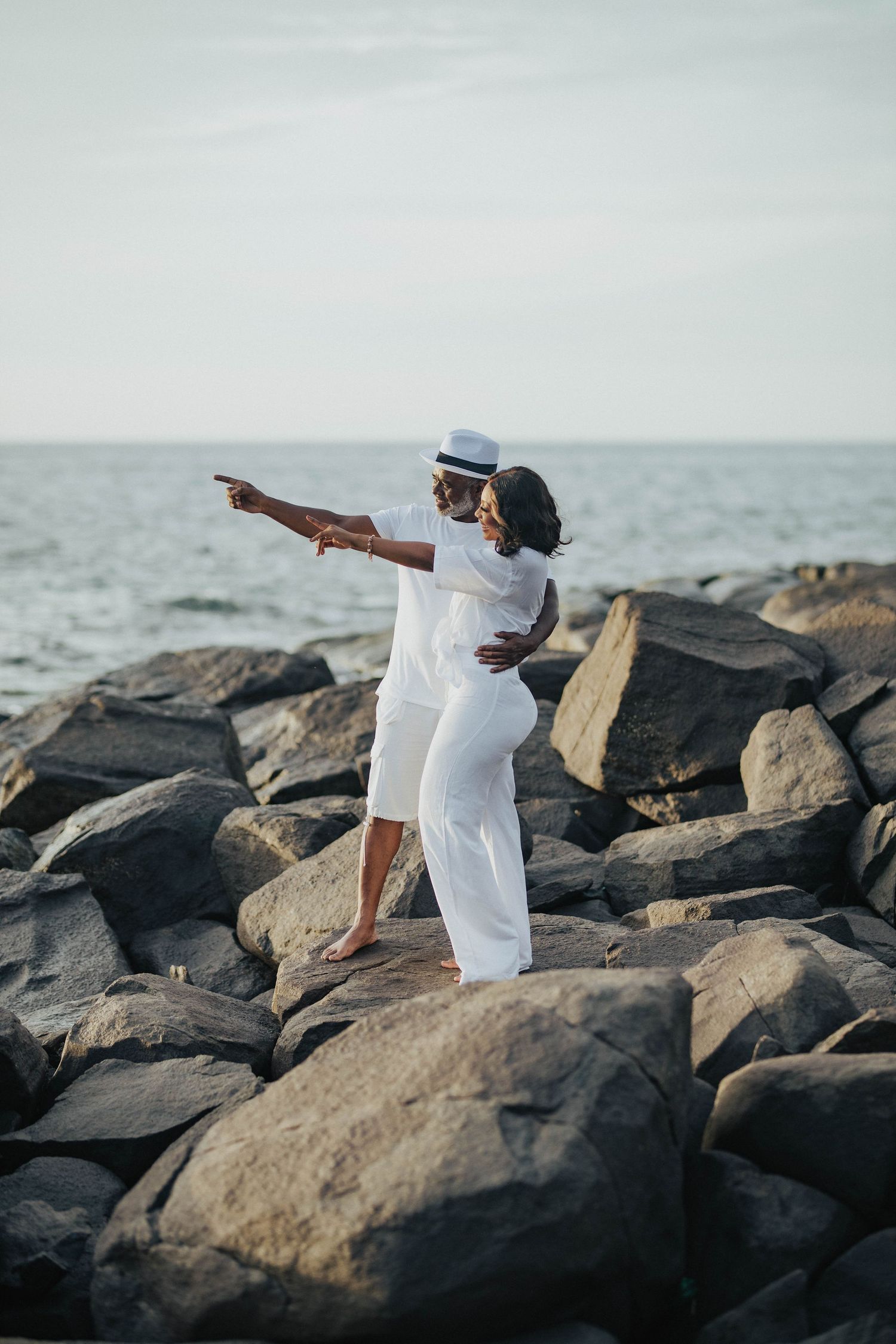 A couple in white outfits stands on coastal rocks pointing toward the ocean horizon.
