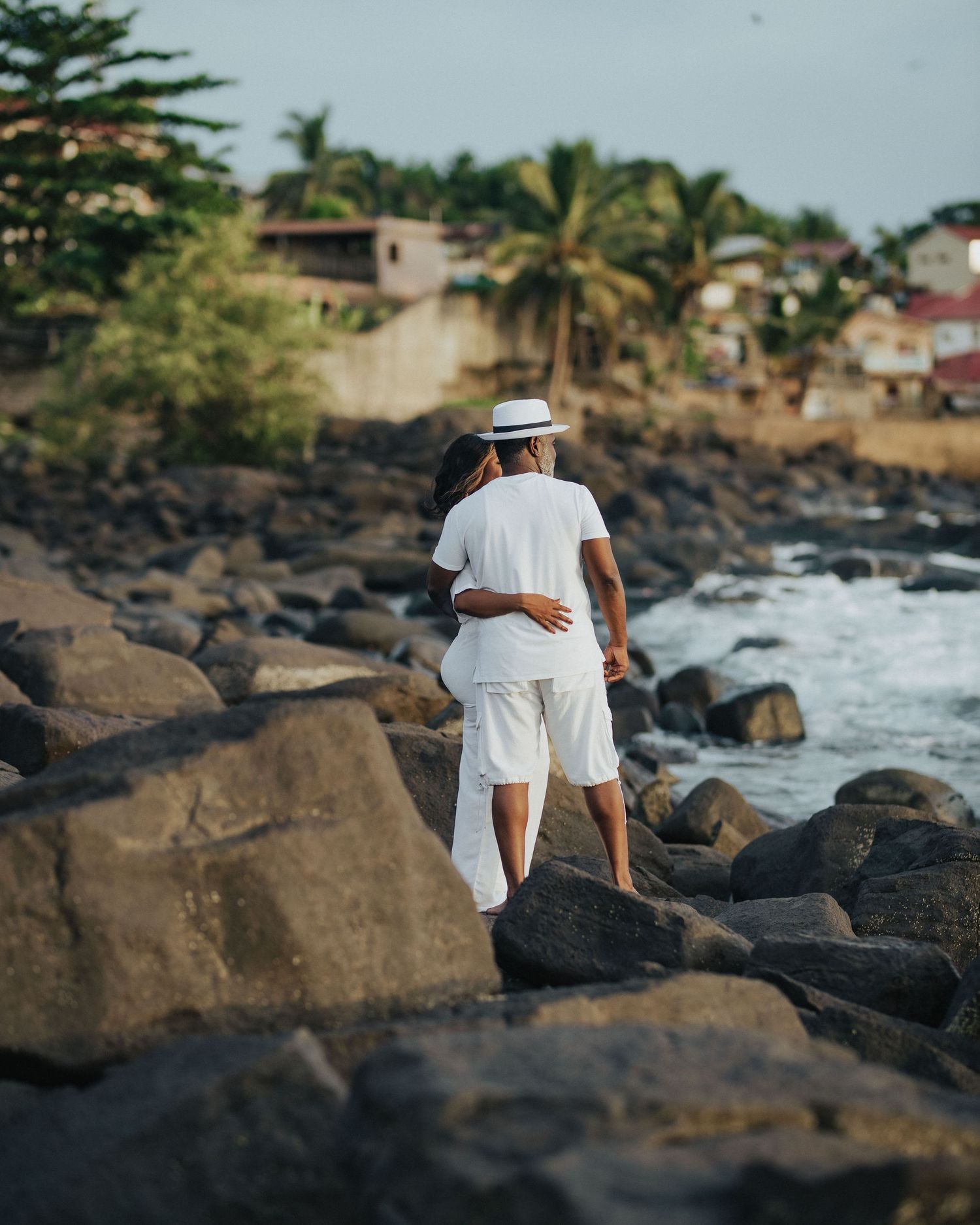 A couple in white outfits walks along rocky coastal shoreline with palm trees and houses in background.