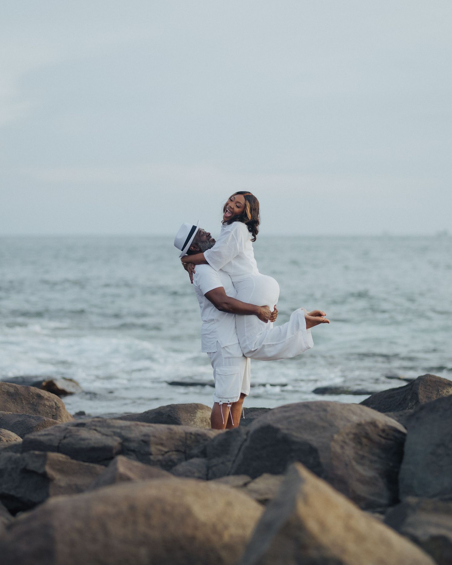 A romantic lift pose of a couple in white clothing on rocky coastal terrain.