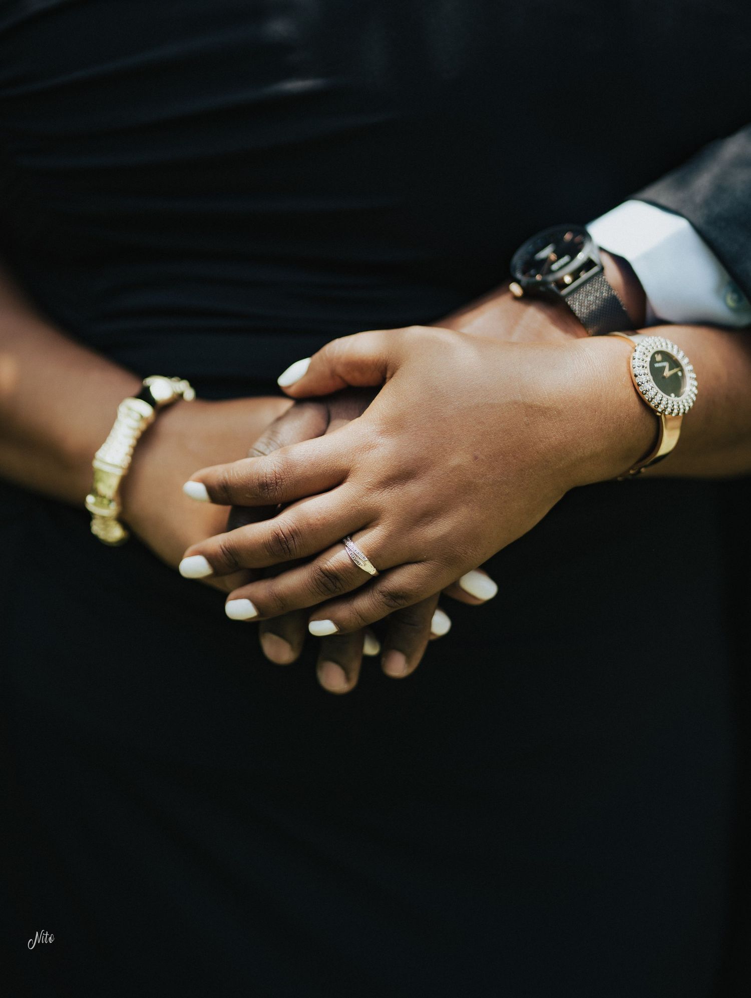 Close-up of clasped hands wearing jewelry and rings against a dark background.