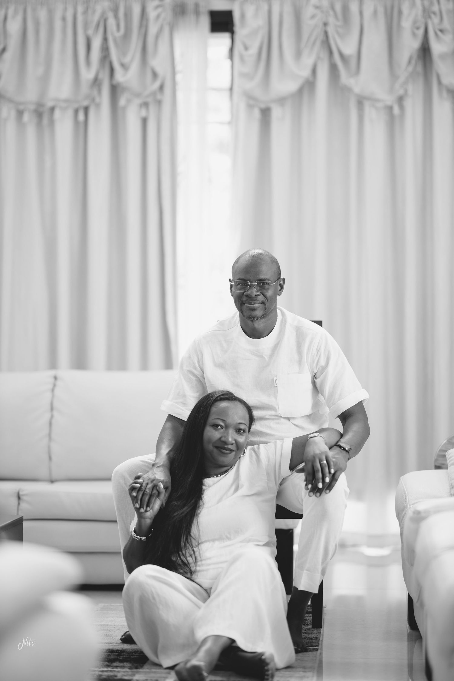 A couple in matching white outfits sharing a casual moment on the floor by a white couch.