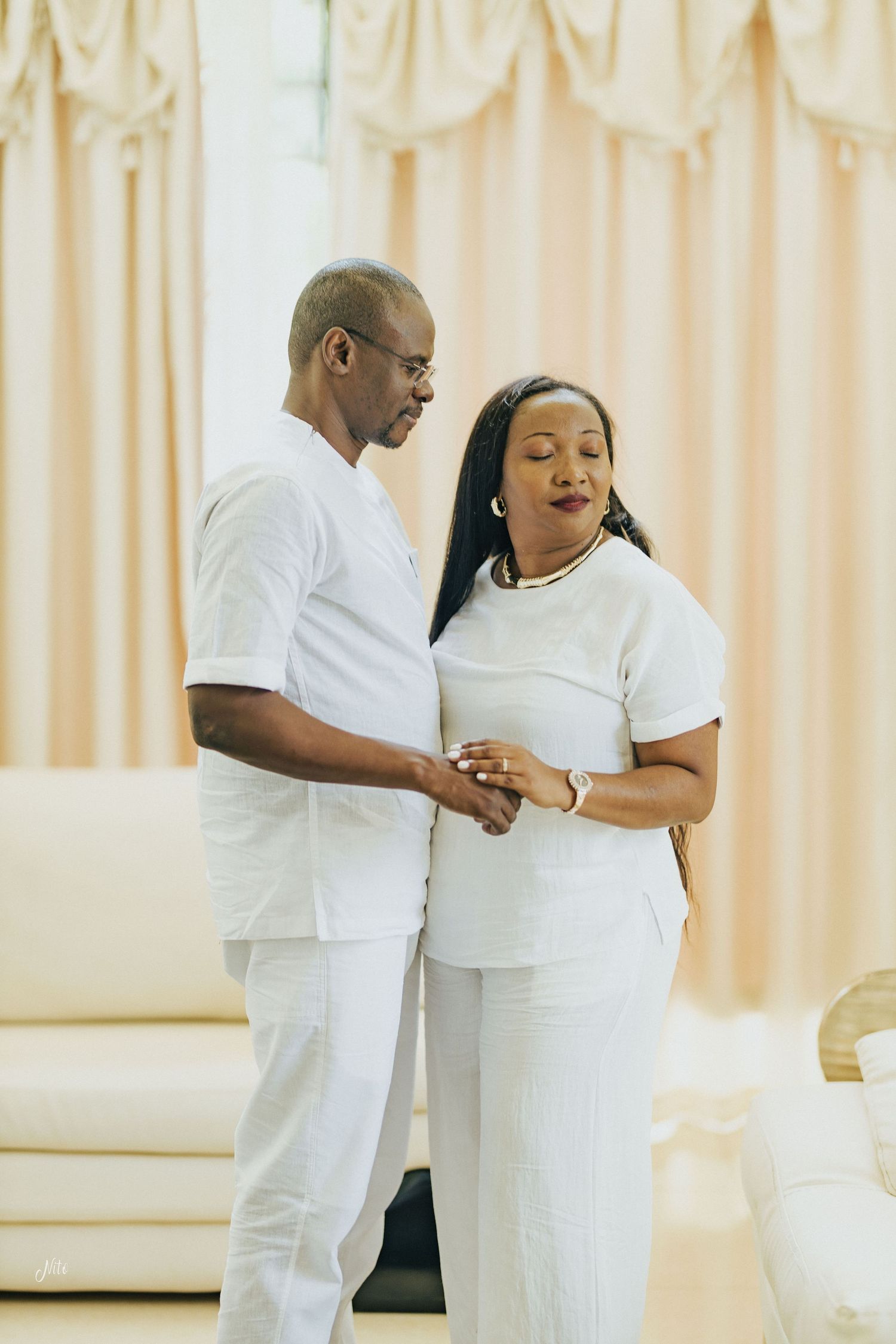 A couple dressed in all white outfits shares an intimate moment together in a bright room with curtains.