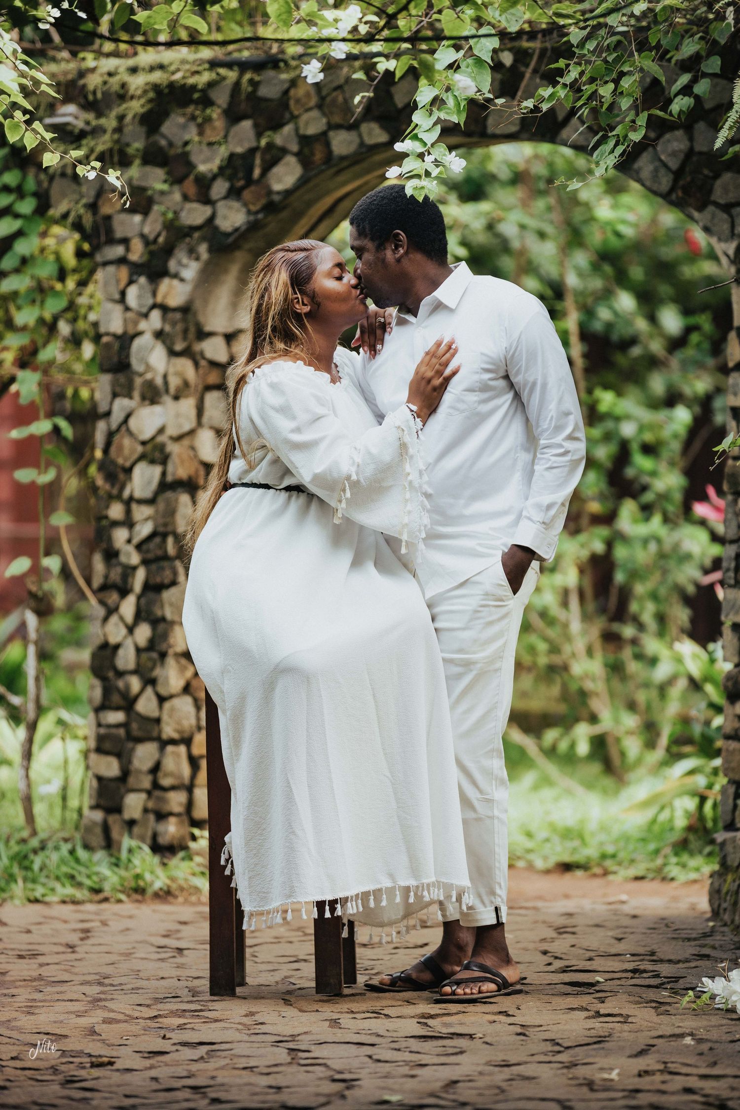 A romantic moment shared between a couple in white attire under a stone archway surrounded by greenery.