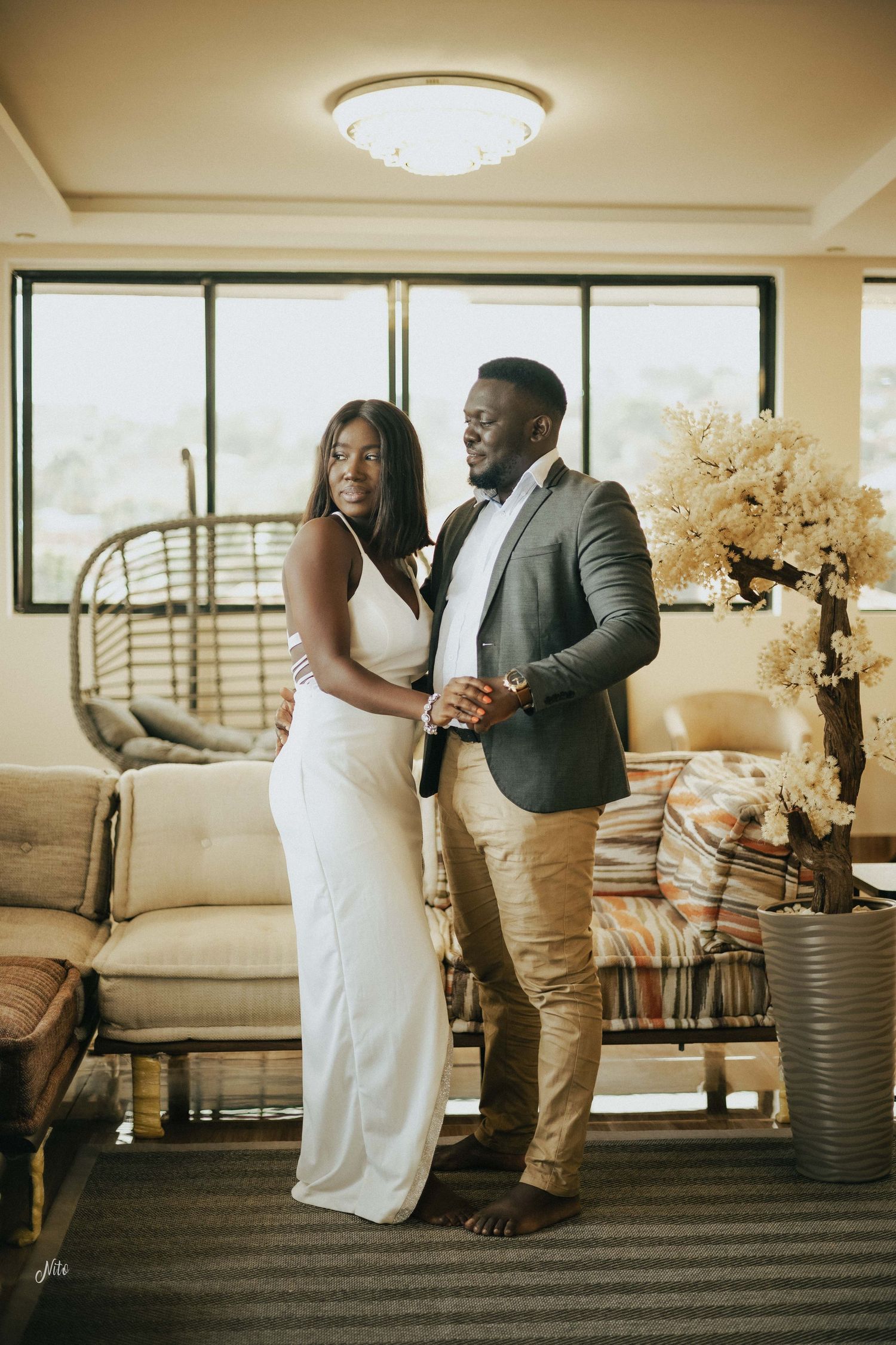 A couple poses together in formal attire near large windows and modern decor.