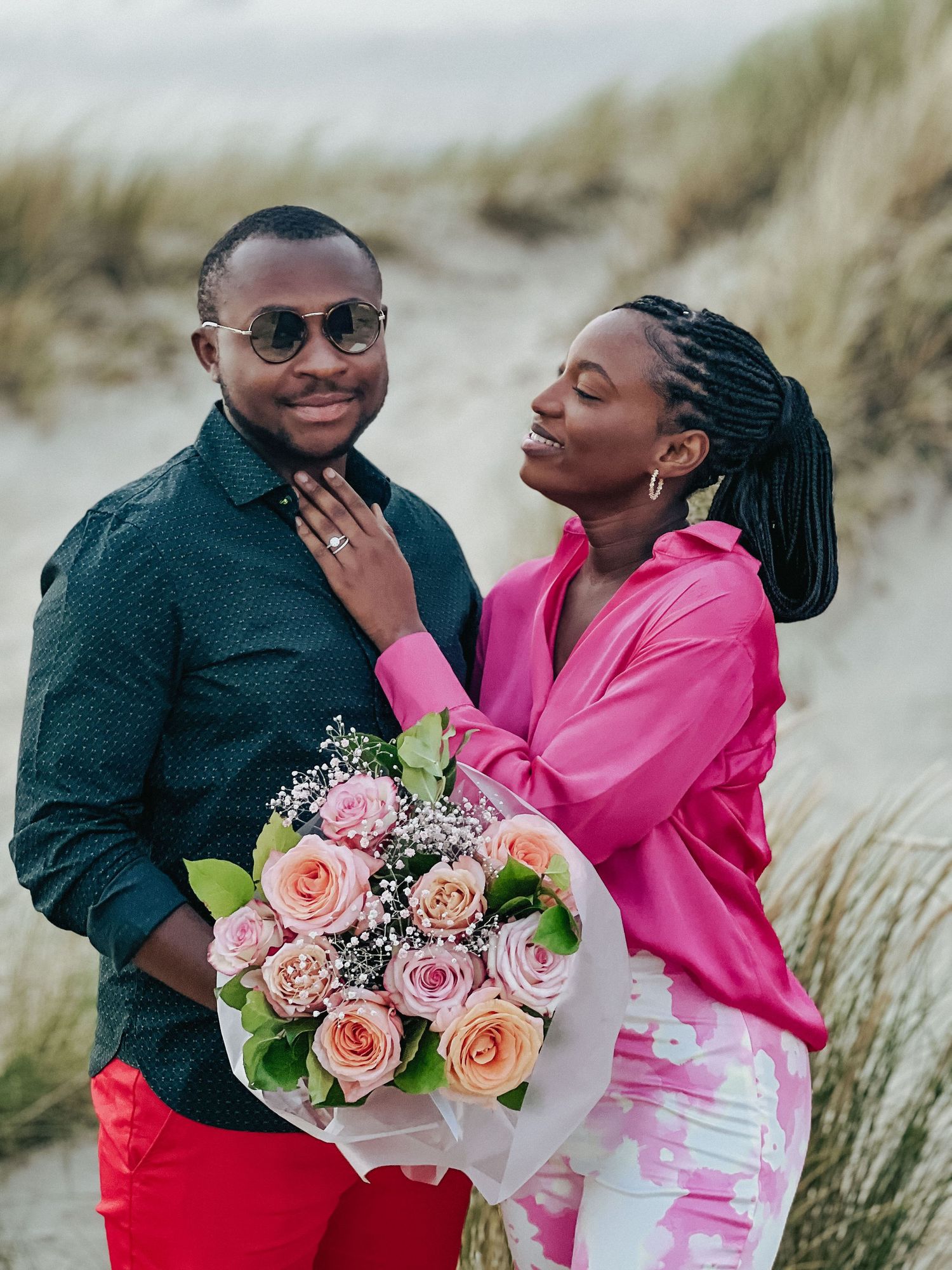 A couple in casual attire sharing a romantic moment with pink roses against beach grass.