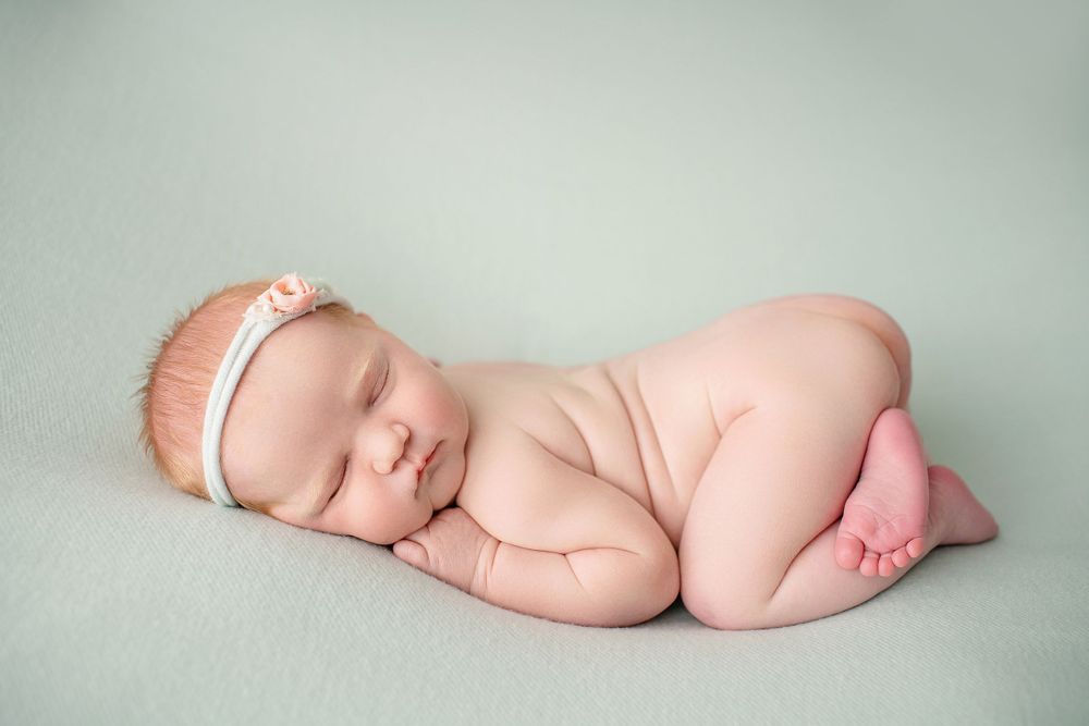 Sleeping newborn baby in curled pose on mint green backdrop with delicate lace headband.