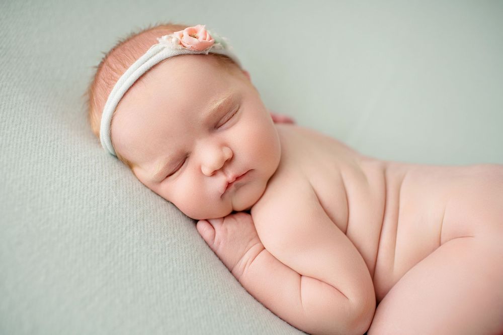 Peaceful newborn resting on side with peach headband against mint green background.