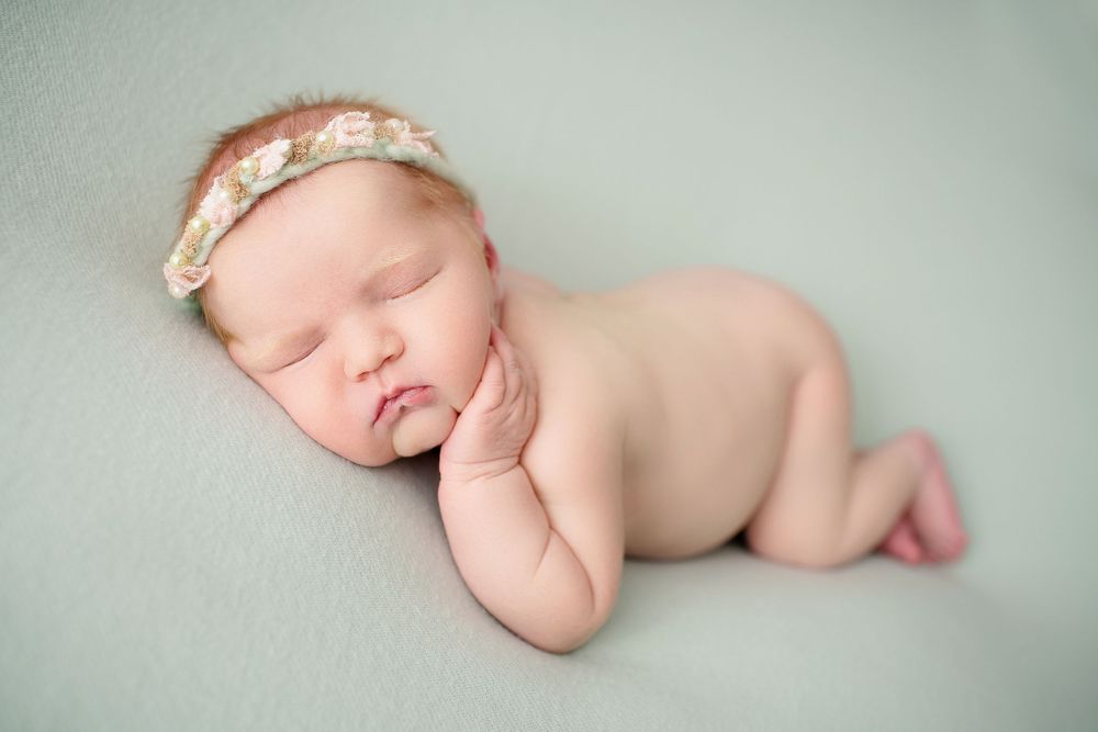 Newborn wearing floral headband sleeping in fetal position on mint green surface.