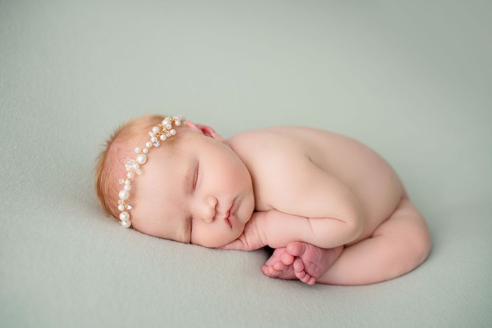 Sleeping newborn with pearl headband curled up on mint green backdrop.