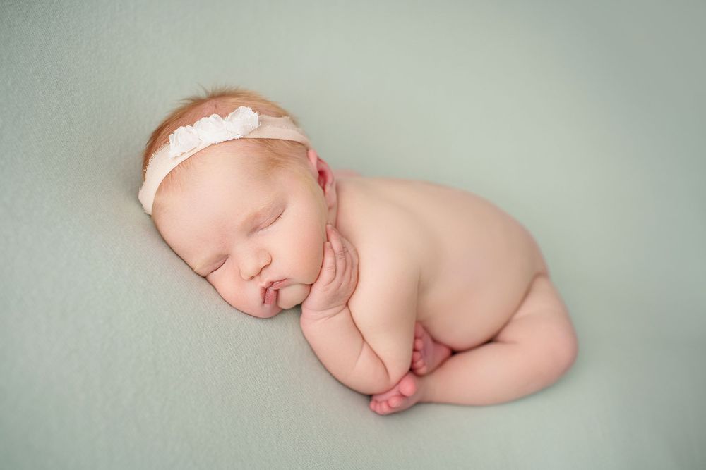 Sleeping newborn with lace headband in curled position on mint background.