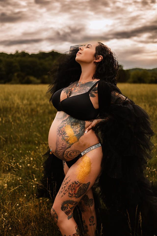 Pregnant woman with tattoos in black outfit posing in field under dramatic sky.