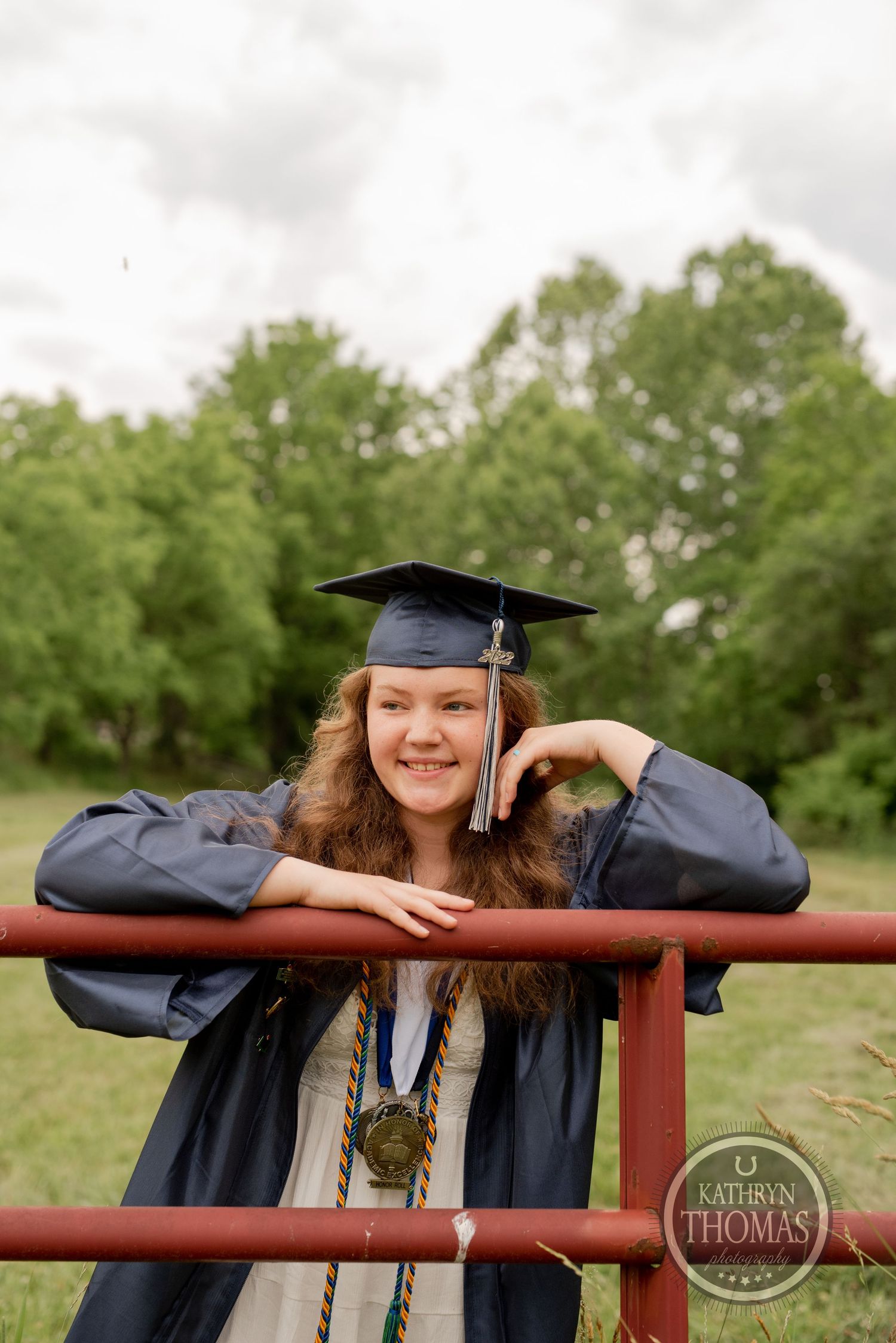 Graduate in cap and gown poses playfully while leaning on red metal railing.