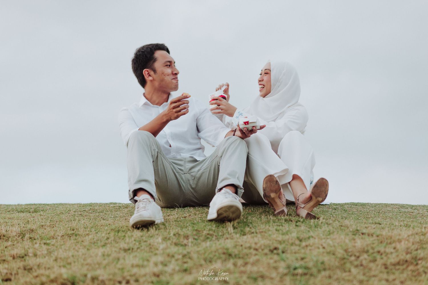 A couple enjoys refreshments together while relaxing on a grassy hillside.