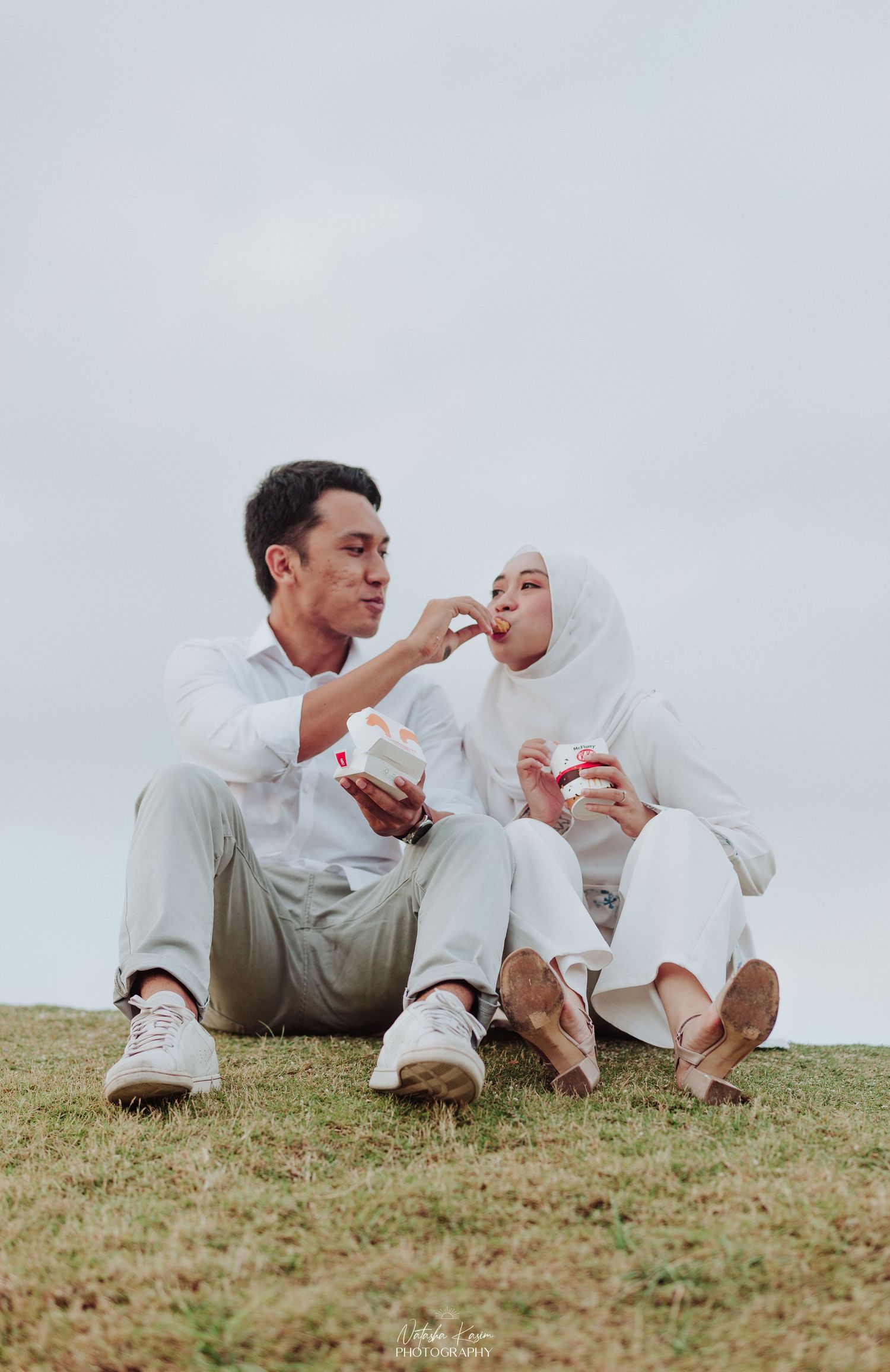 A couple shares snacks while sitting on grass during an outdoor date.