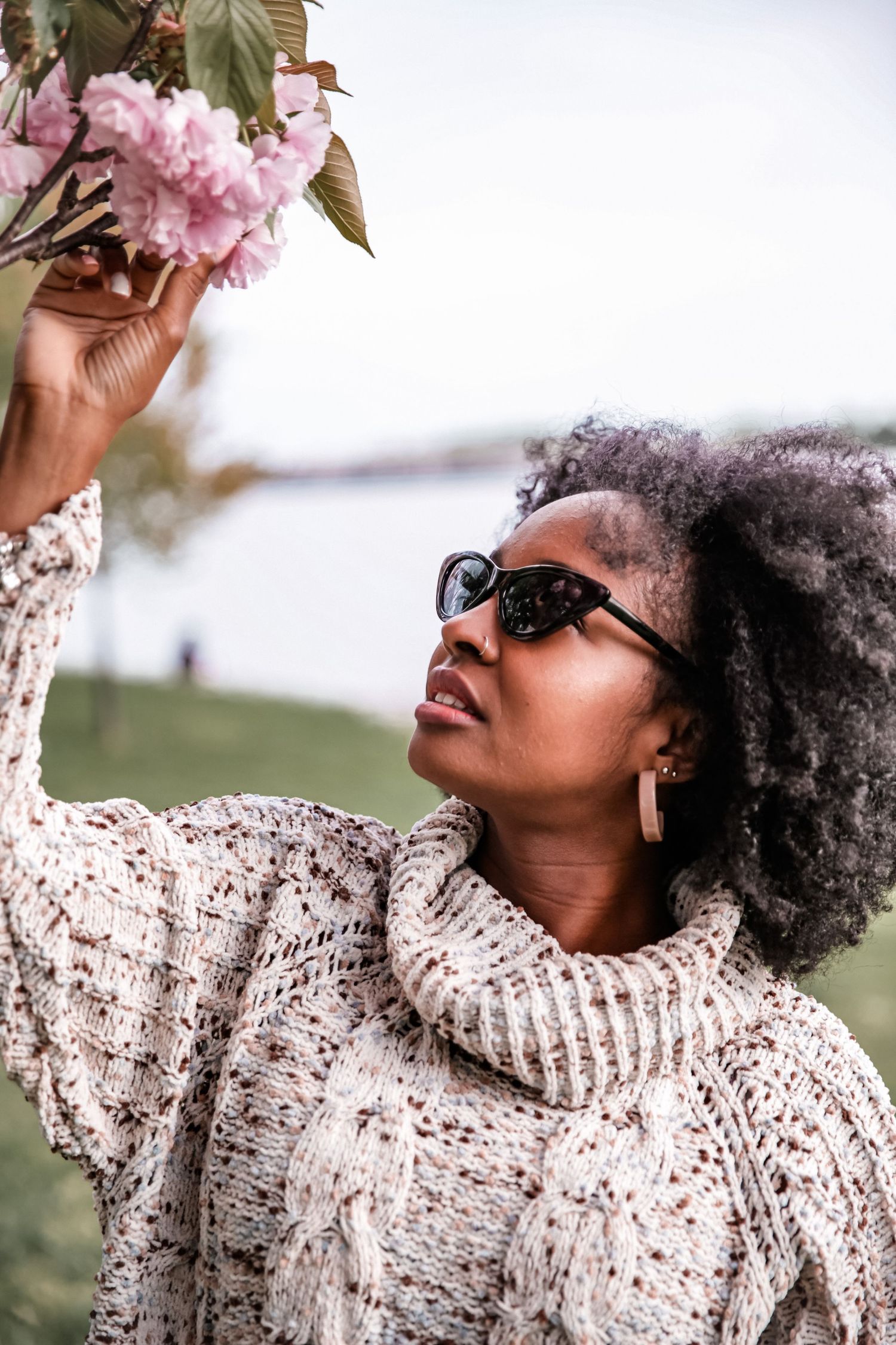 Black woman in sunglasses and knit turtleneck sweater reaching up to touch pink cherry blossom flowers outdoors.