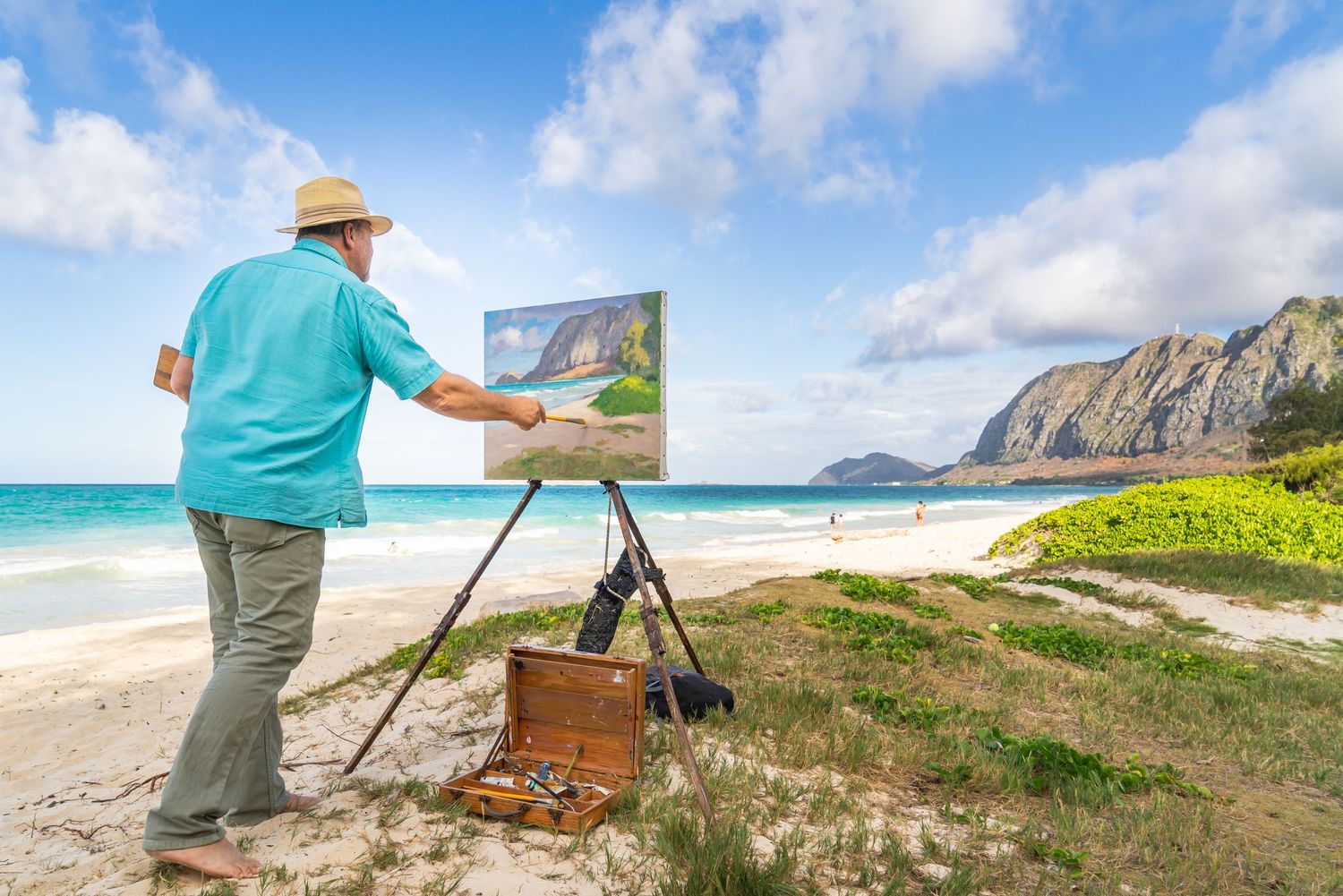 Artist in blue shirt paints coastal landscape on canvas at scenic Hawaiian beach.