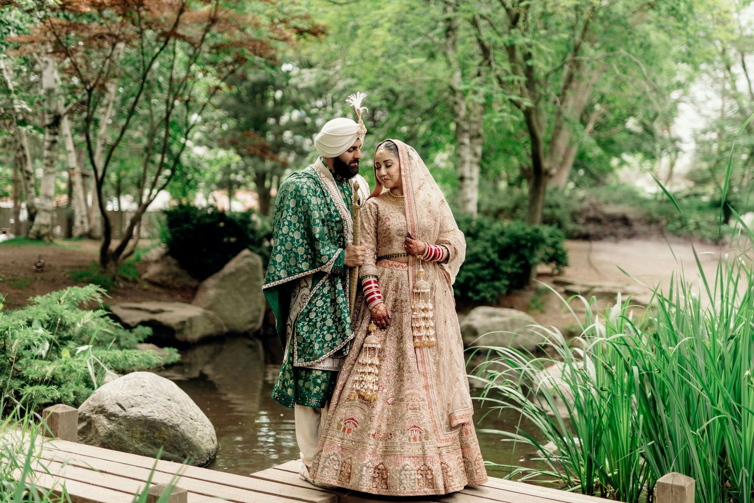 A couple in traditional Indian attire poses on a wooden bridge in a garden setting.