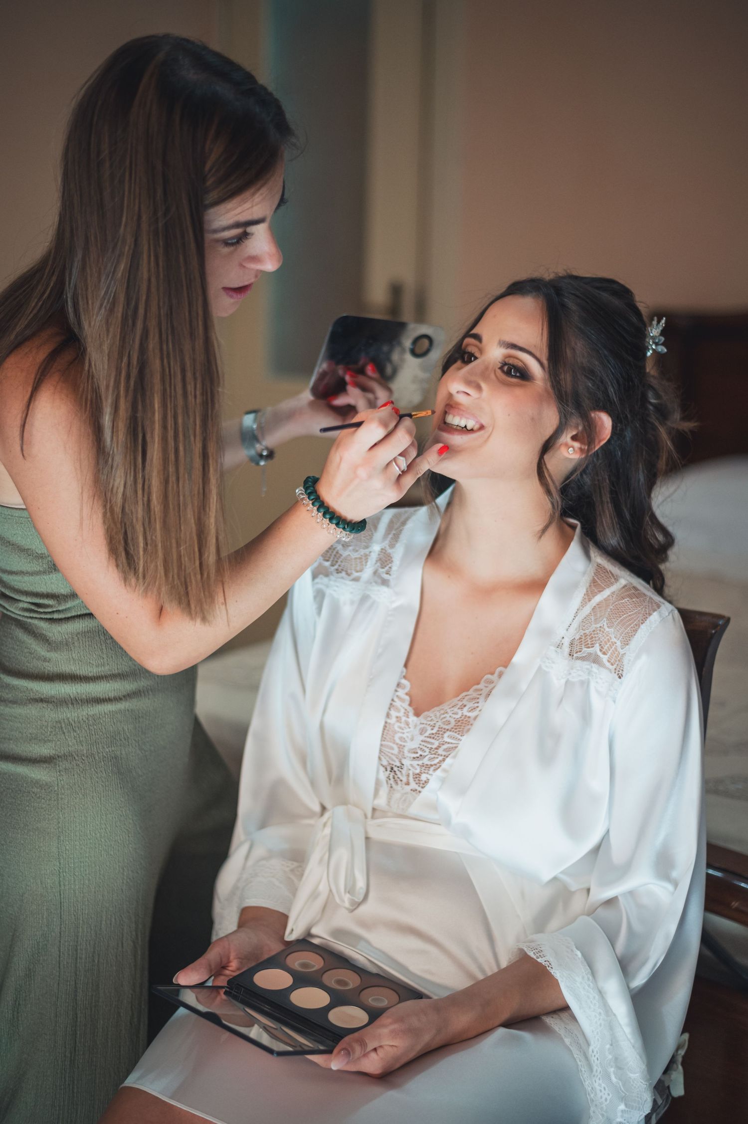 Makeup artist applies lip color while client holds eyeshadow palette in white robe.