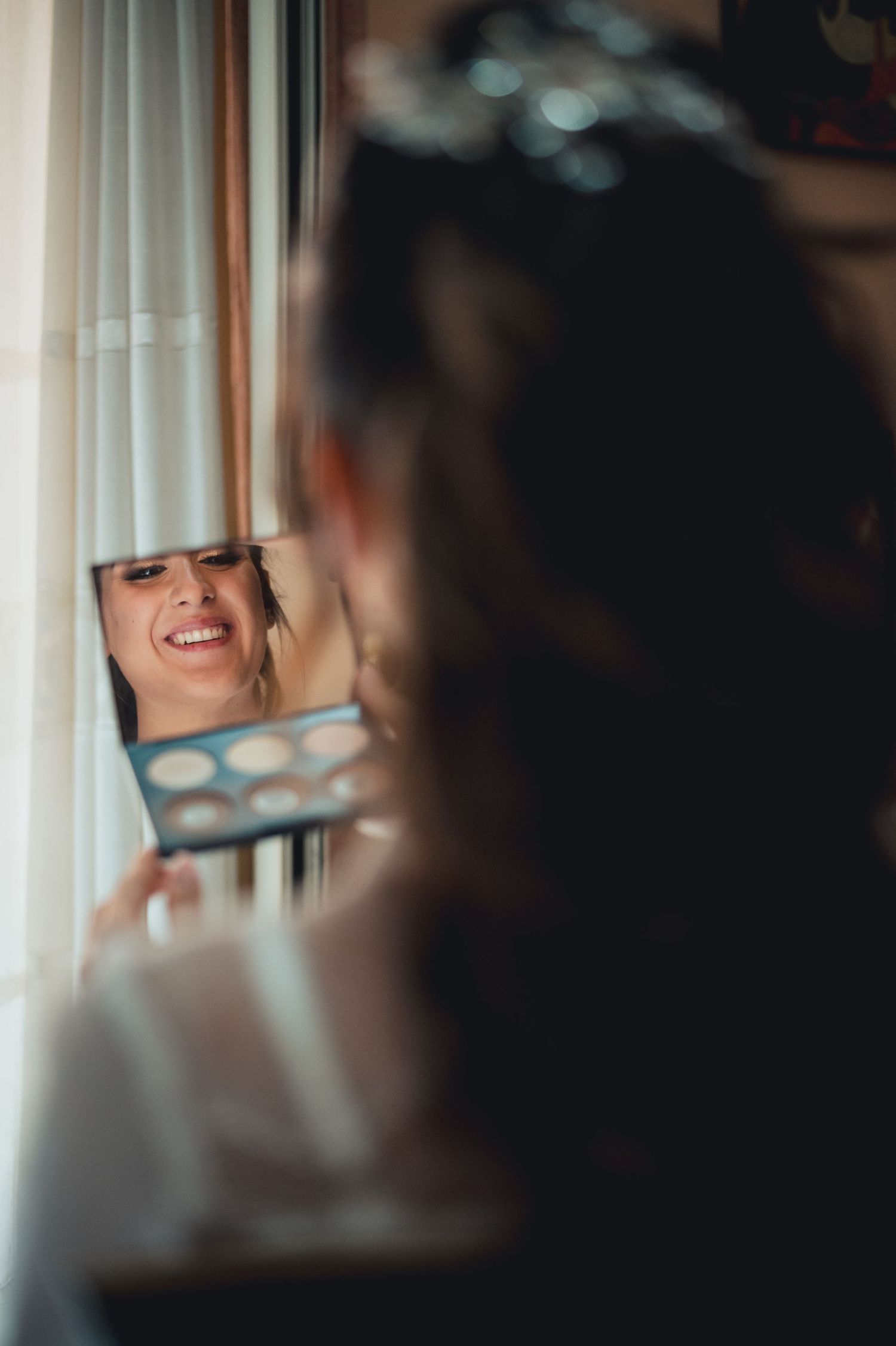A reflection in a handheld mirror shows a smiling expression during makeup preparation.