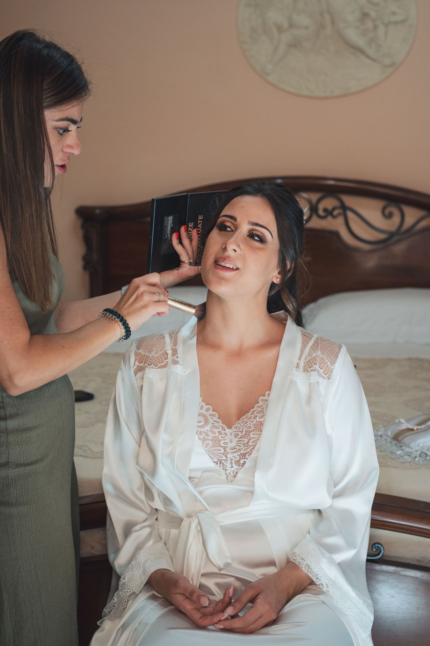 A makeup artist applies cosmetics to someone wearing a white lace robe while seated on a bed.