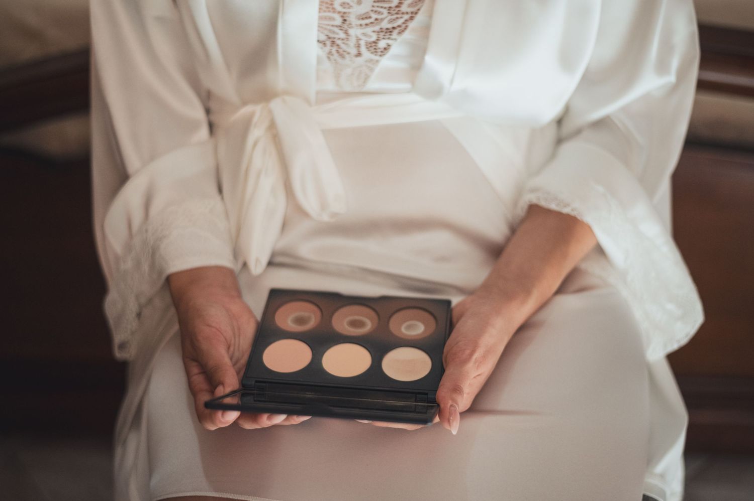 Hands hold a neutral toned eyeshadow palette while wearing a white silk robe.