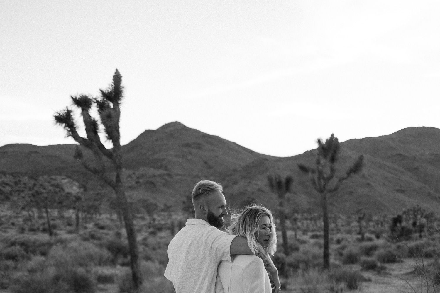 Couple stands together in desert landscape with Joshua trees silhouetted against mountains.