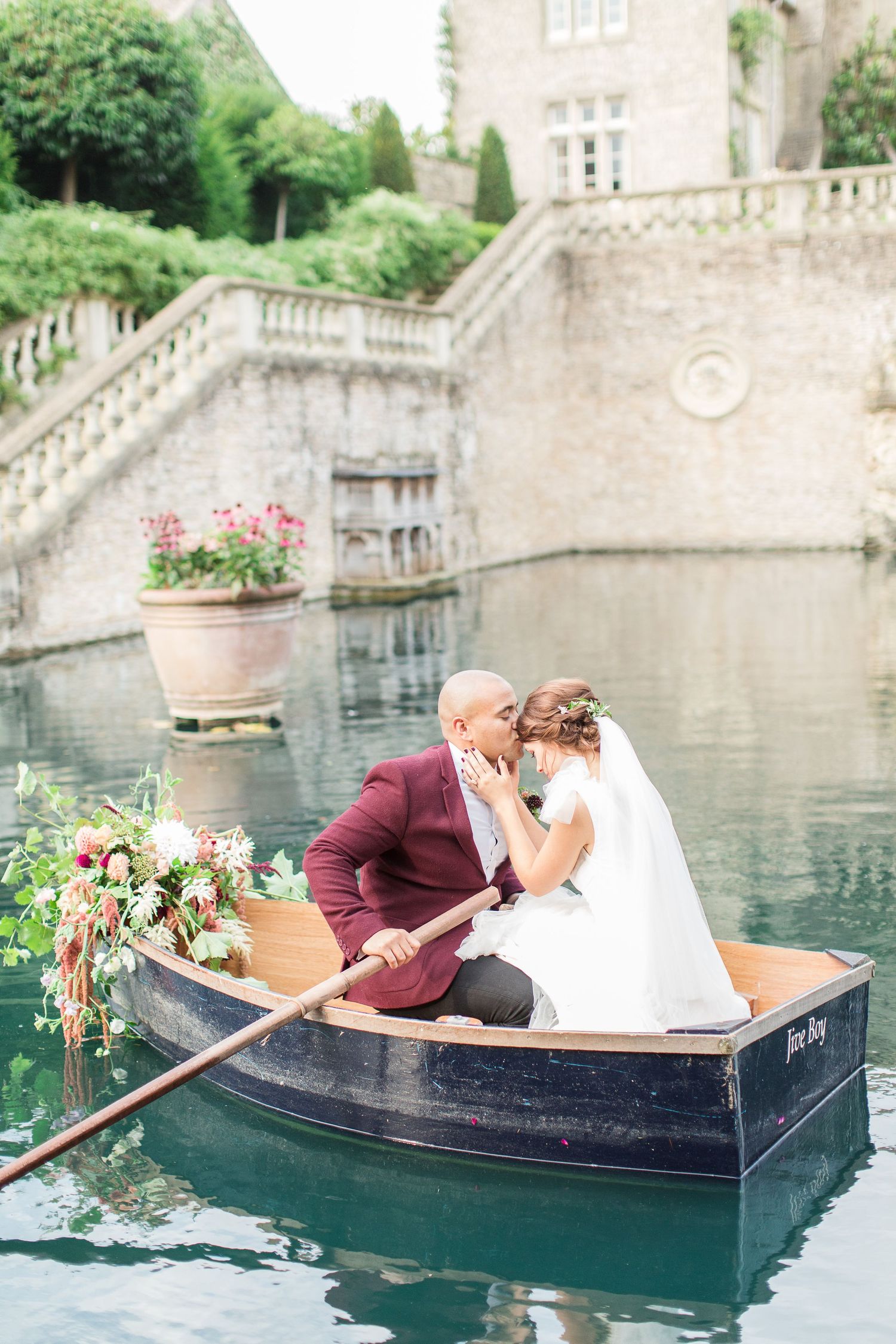Two figures share a romantic golden hour moment in a vintage rowboat on a serene castle pond surrounded by stone architecture euridge manor