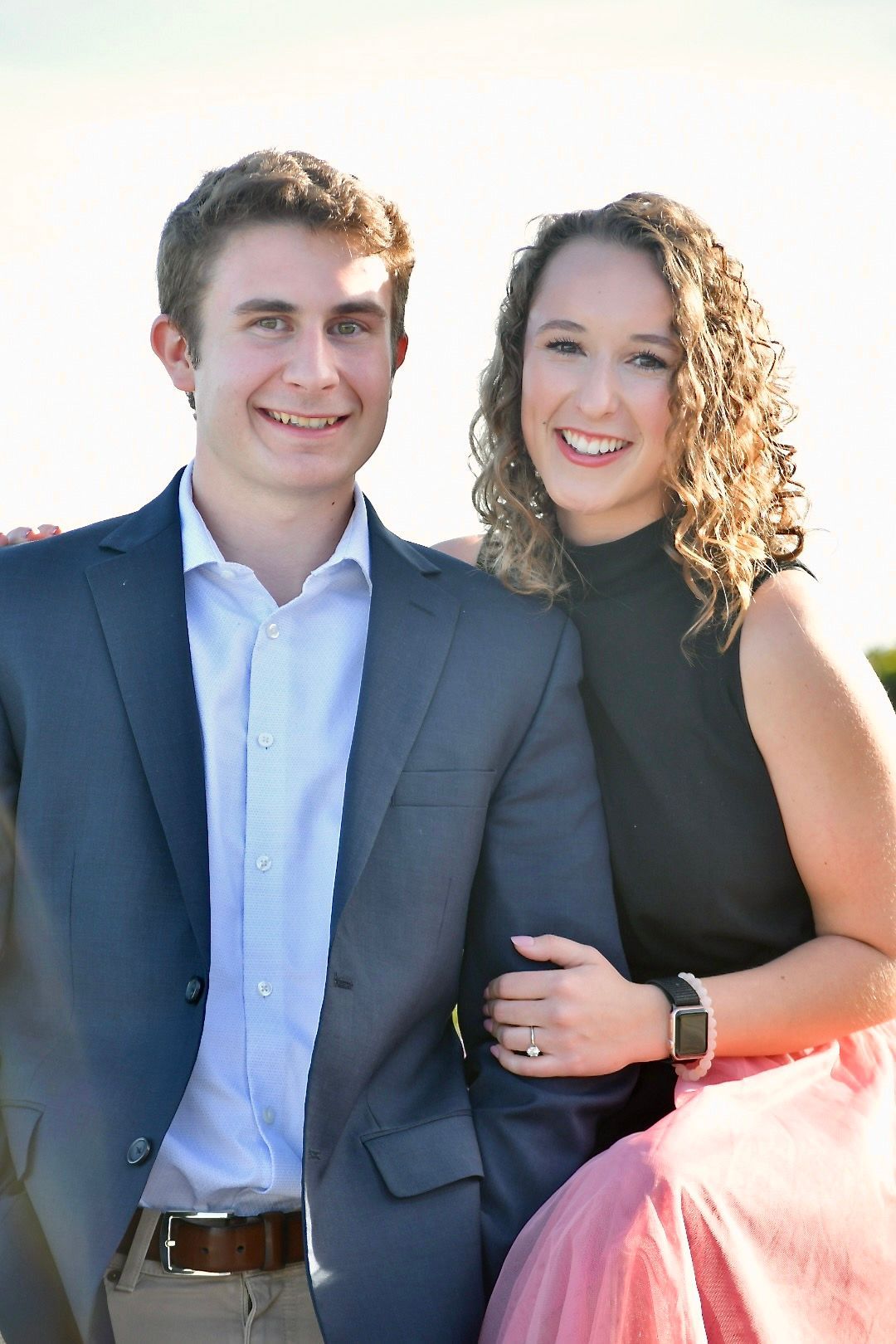 A couple dressed in formal attire pose together outdoors, with the woman wearing a black sleeveless top and the man in a navy suit.