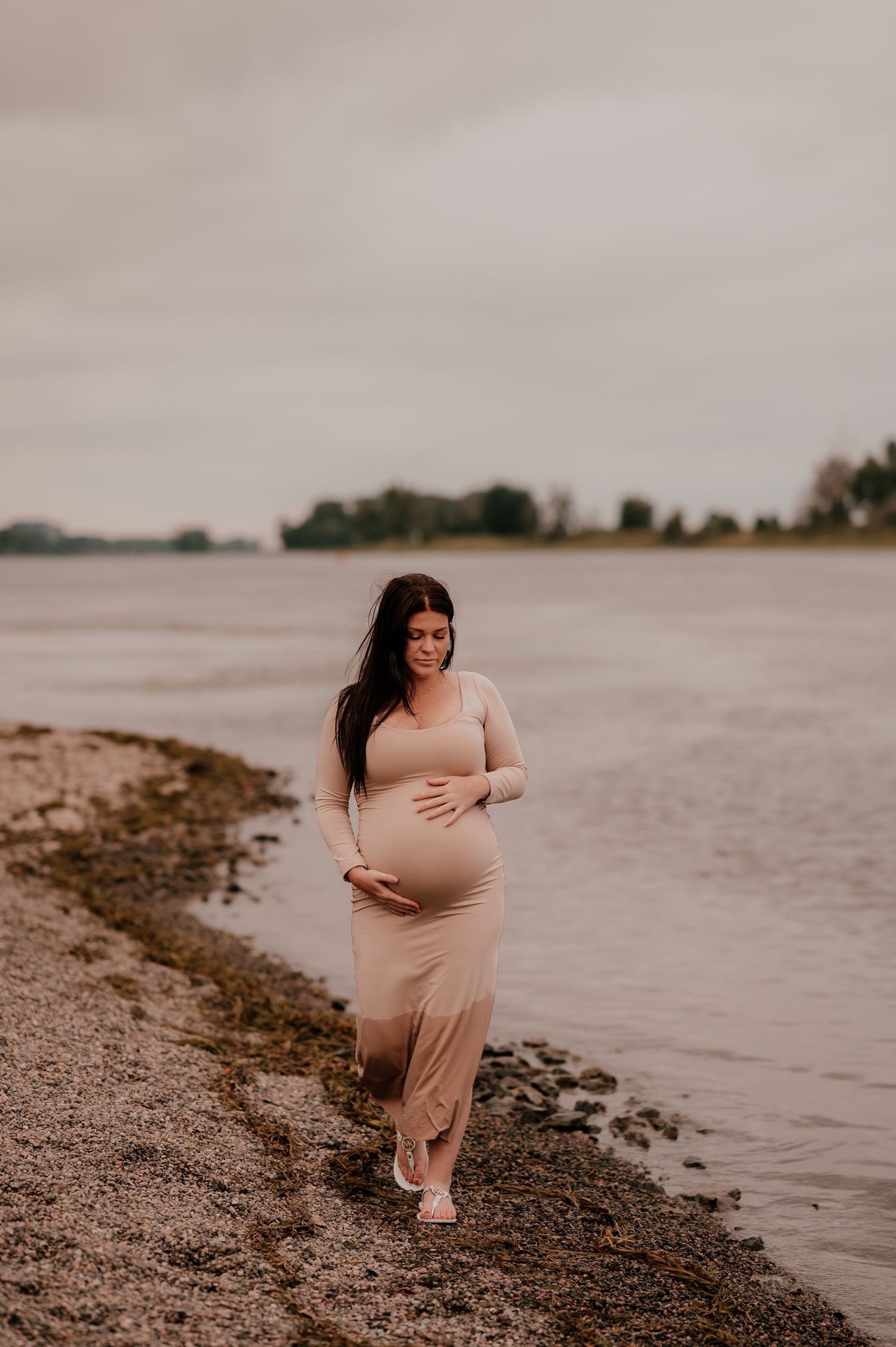 A pregnant woman in a beige dress walks along a misty shoreline.