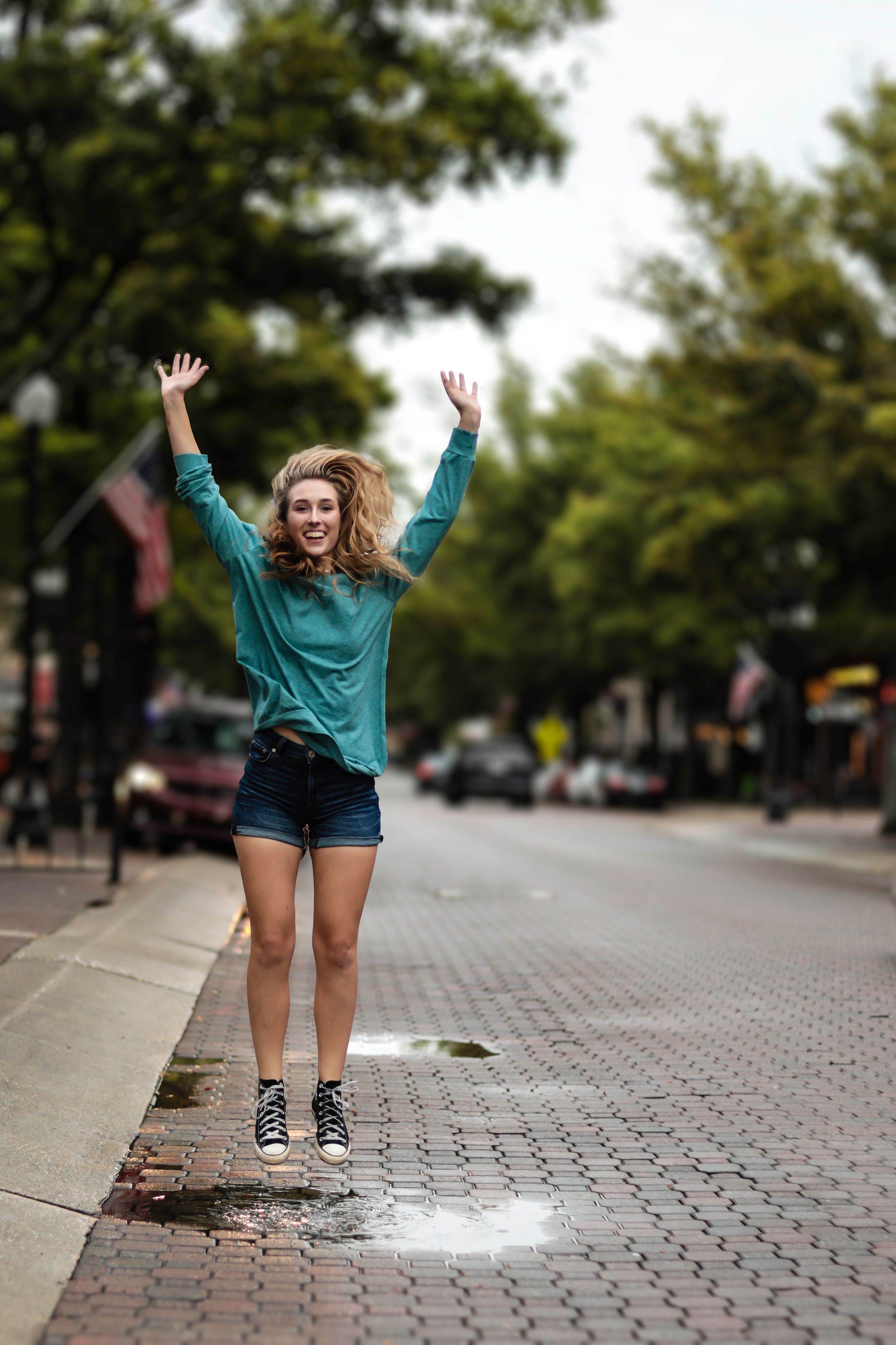 Joyful jumping pose on a brick sidewalk wearing teal sweater and denim shorts.