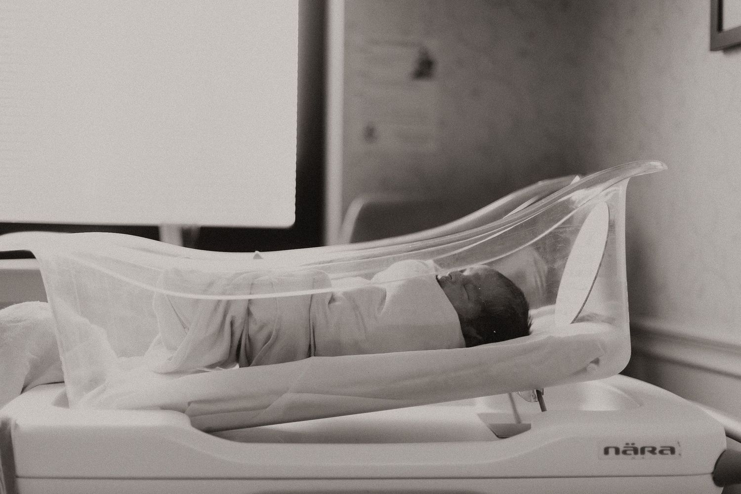 Newborn baby sleeping under protective clear hospital bassinet cover.
