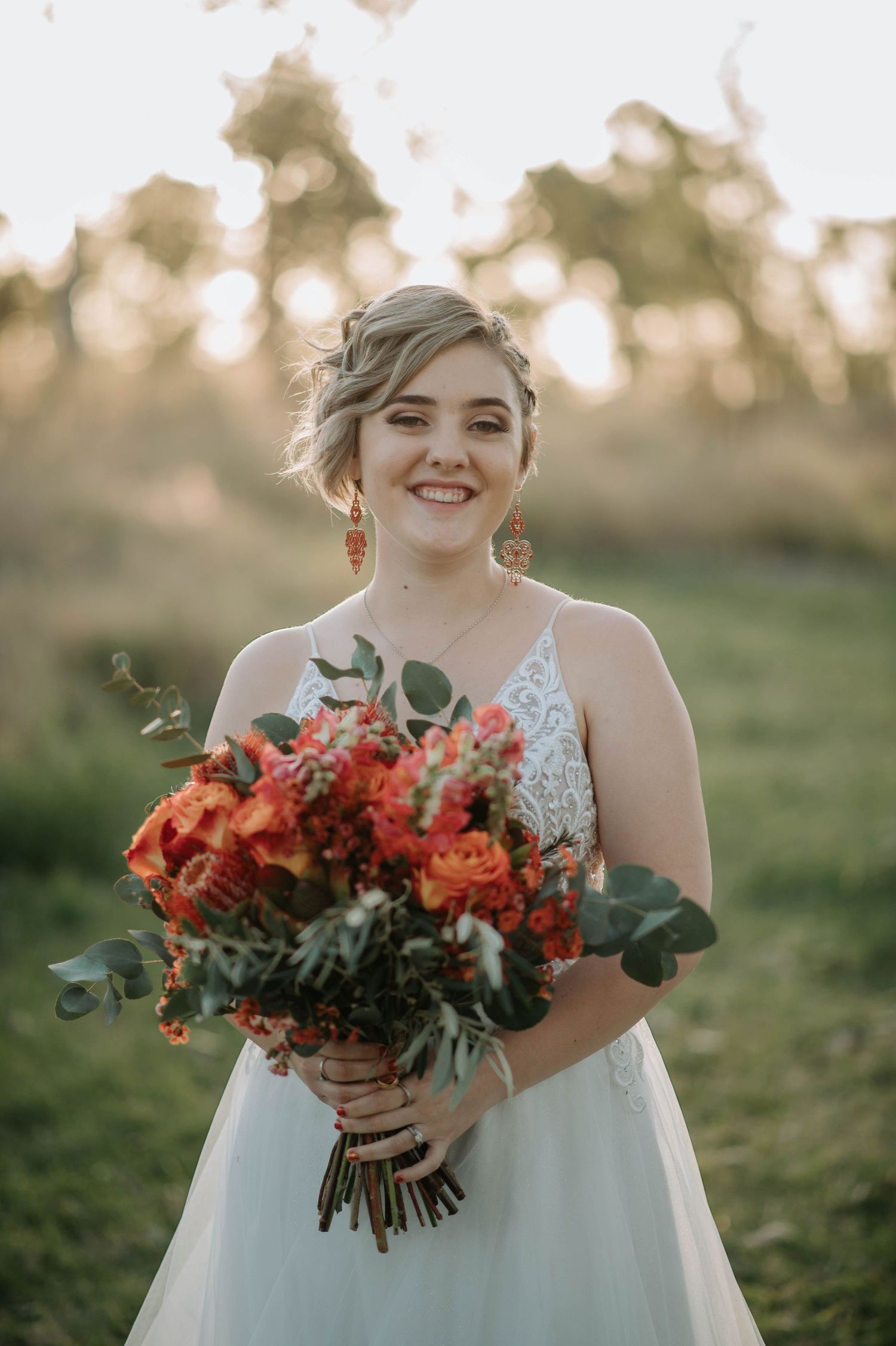 Bride holding vibrant orange and red floral bouquet with eucalyptus in outdoor setting.