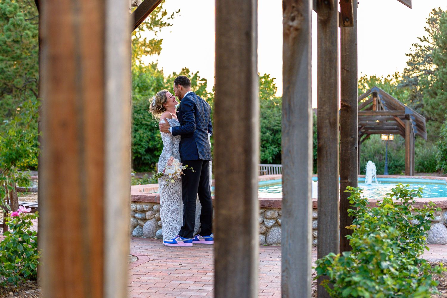 A bride and groom share an intimate moment in a garden in front of a fountain, seen between some posts of an awning.