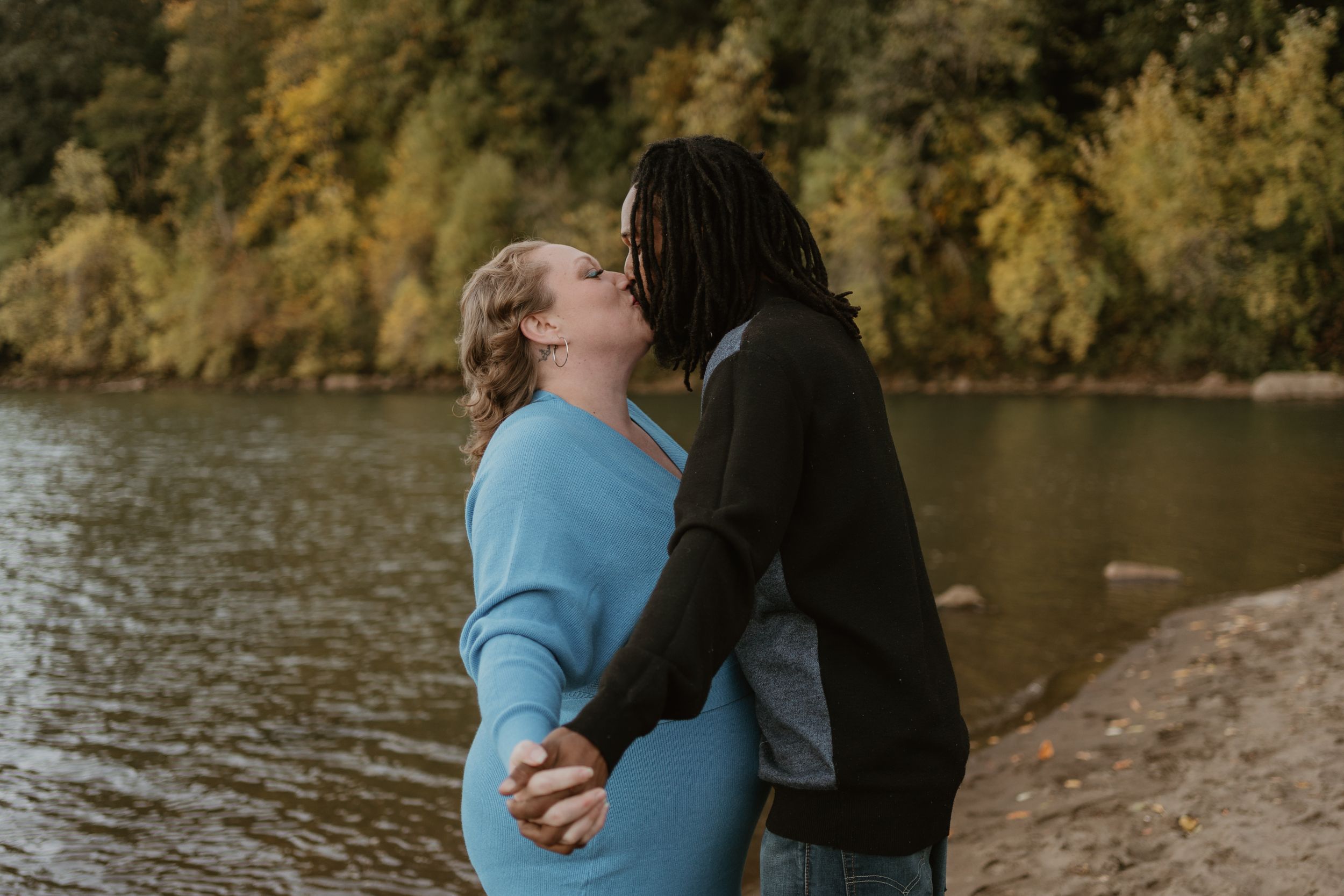 Two people embrace on a sandy shoreline with fall foliage reflecting in the water.