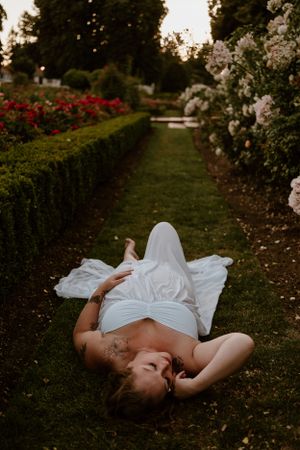 Person in white dress lying on grass surrounded by blooming flowers in a garden setting.