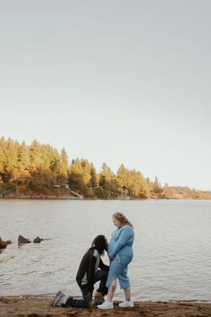 A couple sits together on a rocky beach shore during autumn with golden trees in the background.