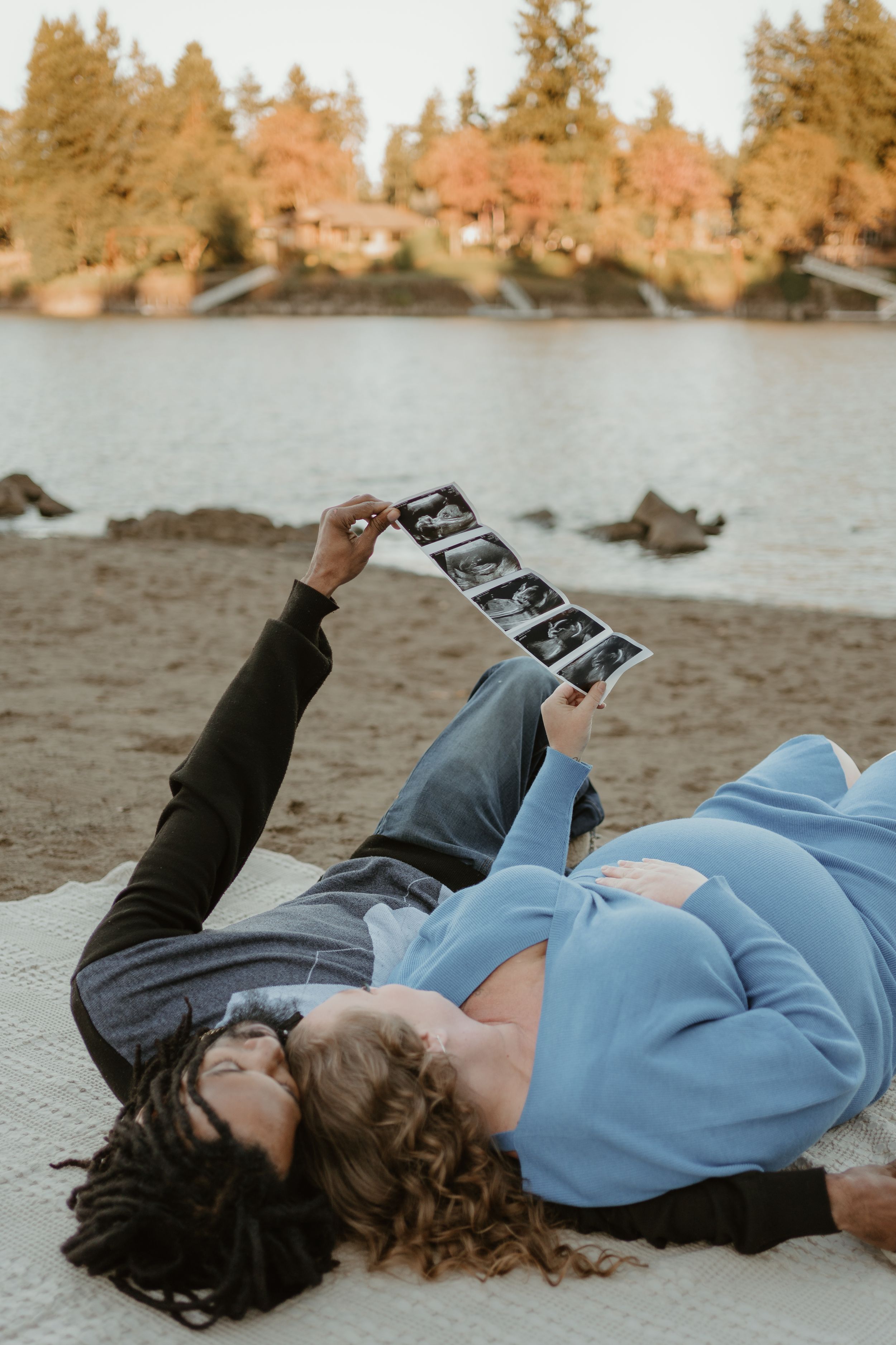 Two people lie on a beach looking at ultrasound photos with fall trees reflecting in the water.
