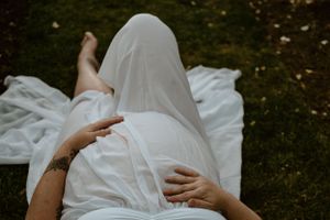 Figure draped in white fabric resting in a garden with blurred background.