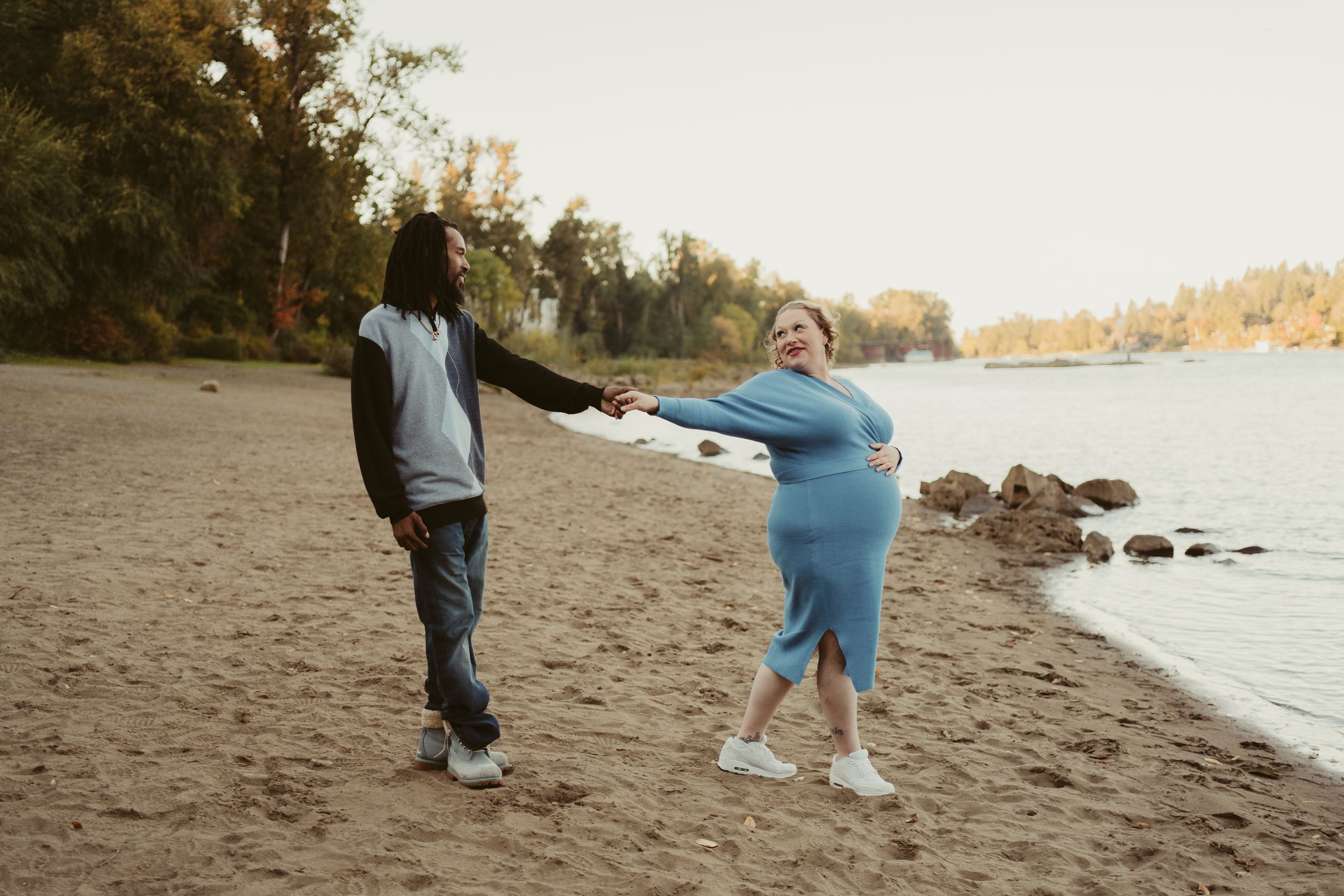 Two people walk along a peaceful beach shore with autumn trees in the background.