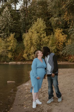 A couple shares an intimate moment on a beach with autumn trees in the background during golden hour.